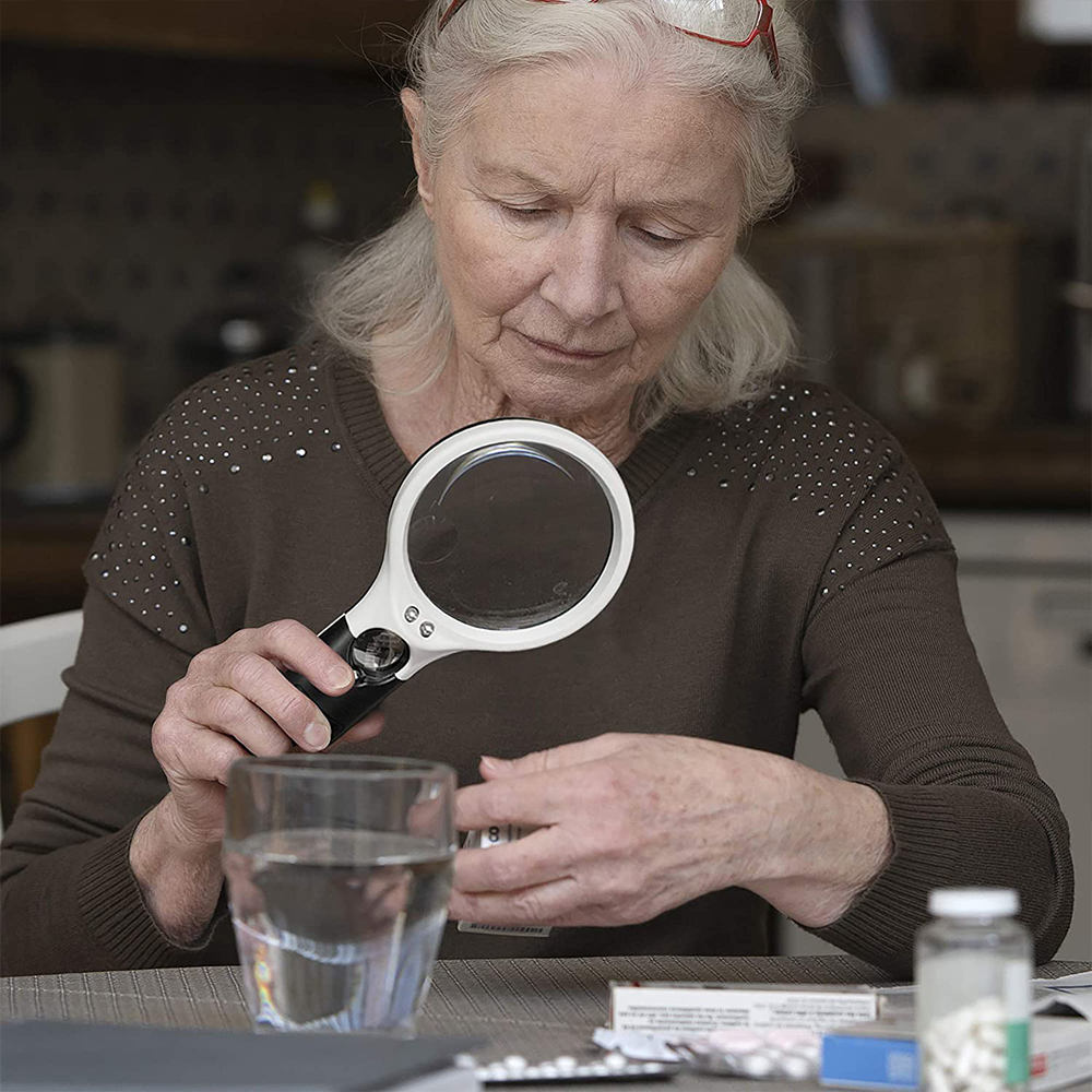 Eine ältere Frau betrachtet eine Pillenflasche mit einer Lupe. Auf dem Tisch stehen ein Glas Wasser und Medikamente.