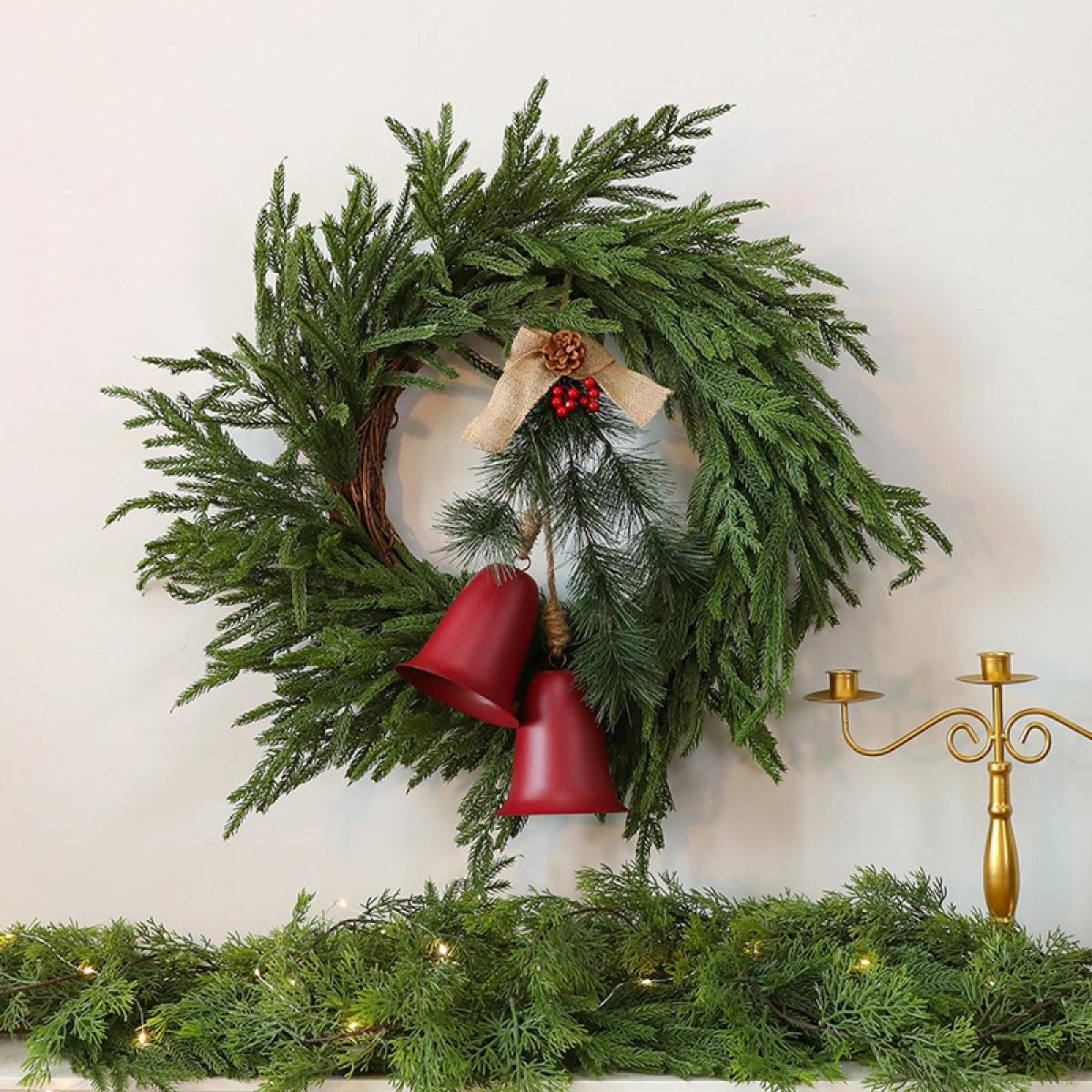 Ein Weihnachtskranz mit roten Glocken, Beeren und einer Schleife hängt an einer weißen Wand. Grüne Girlande und ein goldener Kerzenhalter darunter.