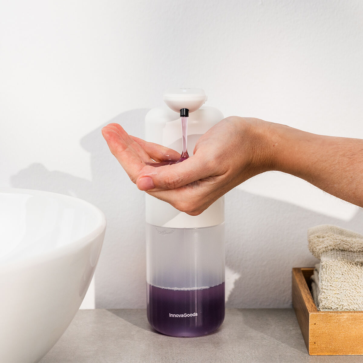 Una mano raccoglie sapone liquido viola erogato da un dispenser automatico bianco. Un lavandino e un asciugamano sono sui lati.