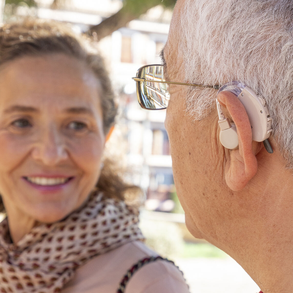 Ein älterer Mann mit Hörgerät und Brille. Eine Frau lächelt im Hintergrund.