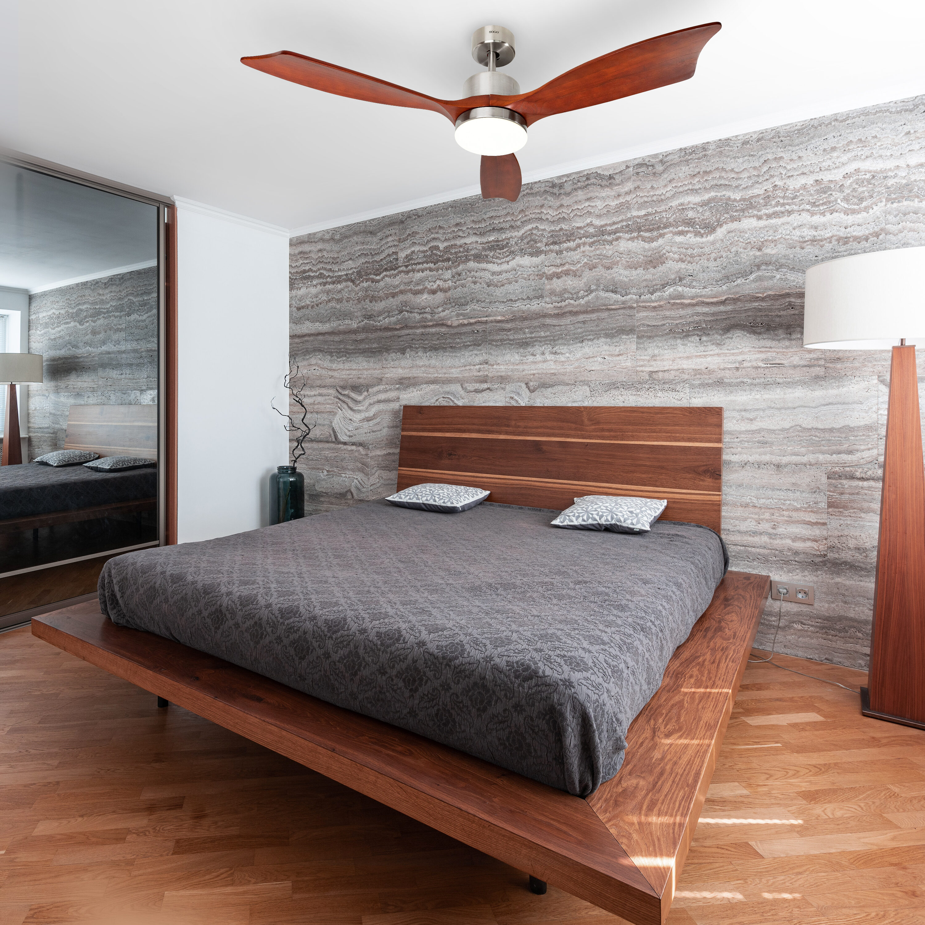 Camera da letto con letto in legno, ventilatore a soffitto e muro in pietra. Visibili anche un grande specchio e una lampada da terra.