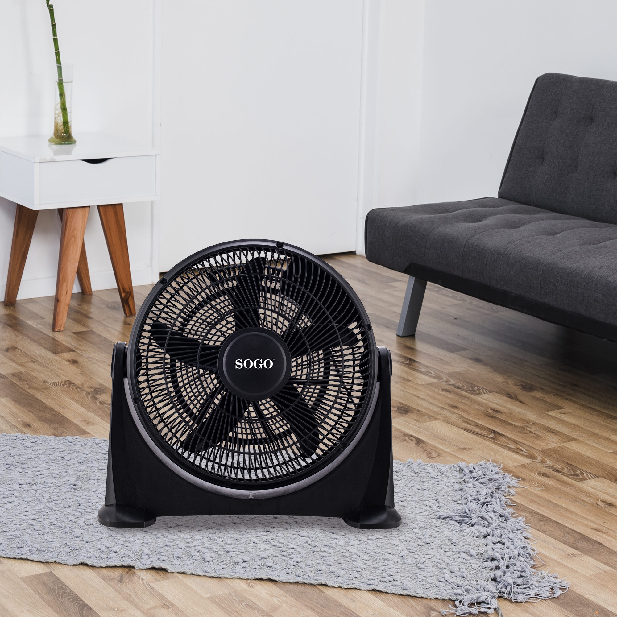 Un ventilador negro SOGO sobre una alfombra gris. A la izquierda, una mesa auxiliar blanca con una planta y a la derecha, un sofá gris.