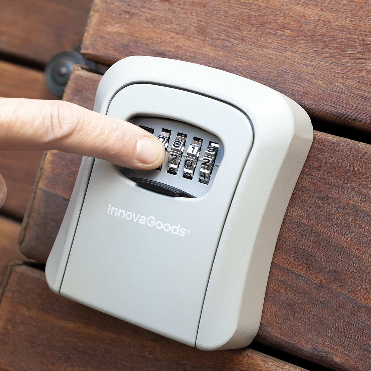 Una mano gira el dial de una caja de cerradura de combinación gris. La caja está sobre tablones de madera. La marca InnovaGoods es visible.