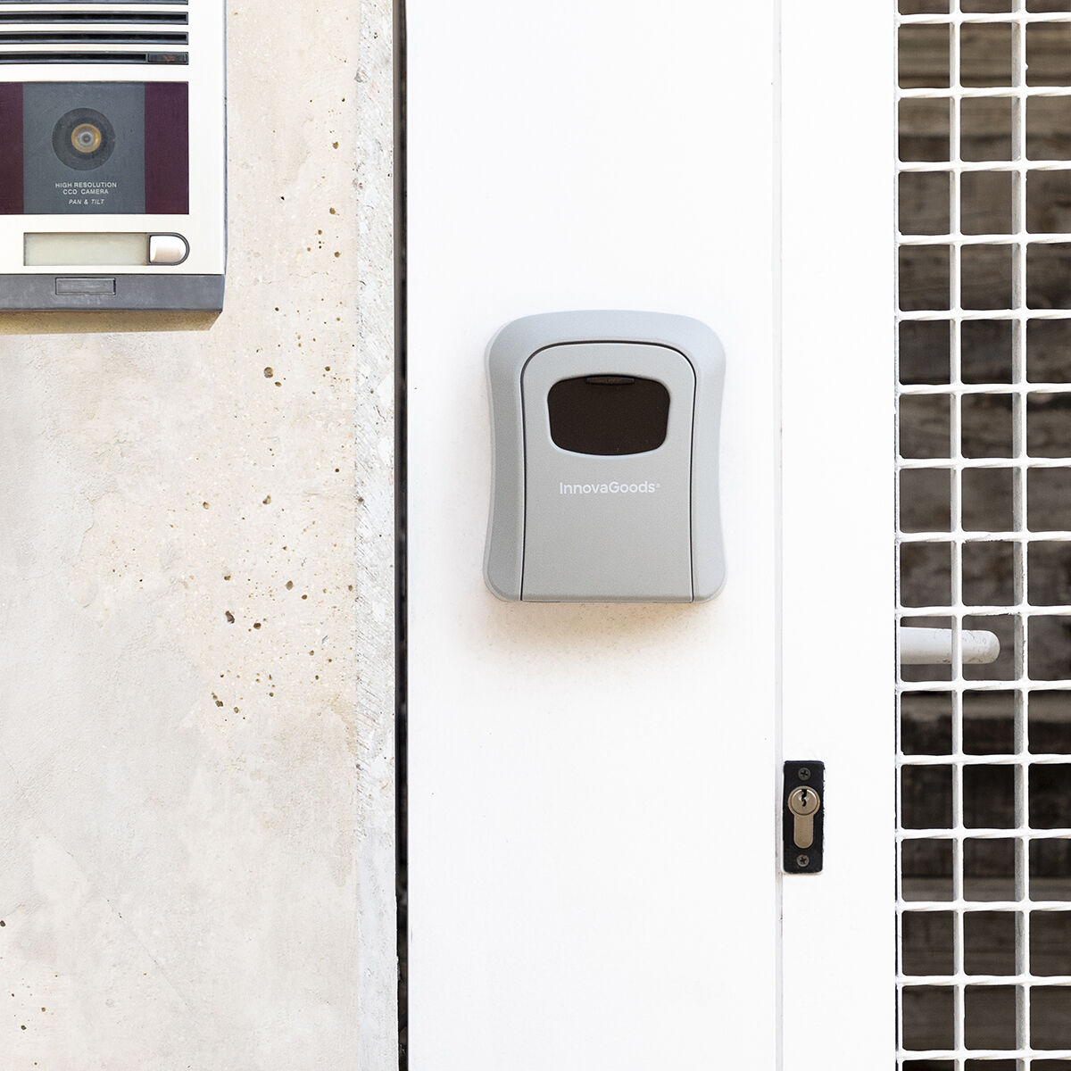 Una caja fuerte gris para llaves está montada en una puerta blanca. Un sistema de intercomunicación blanco está a la izquierda. Una rejilla metálica blanca está a la derecha.