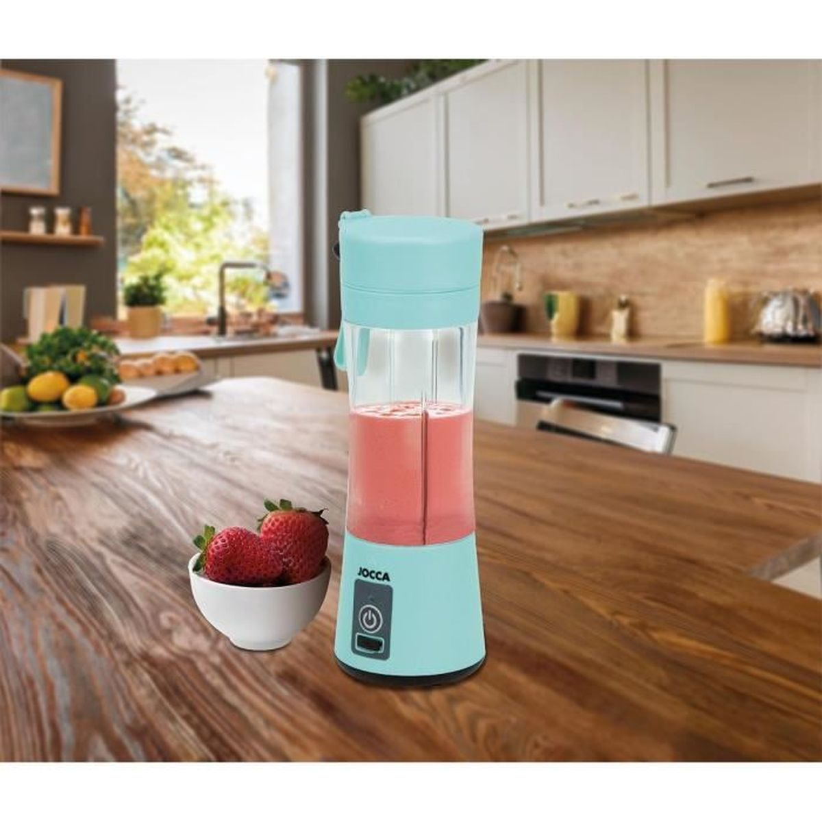 Een turquoise blender met roze vloeistof staat op een houten tafel. Een kom met aardbeien staat in de buurt. De keuken is op de achtergrond.