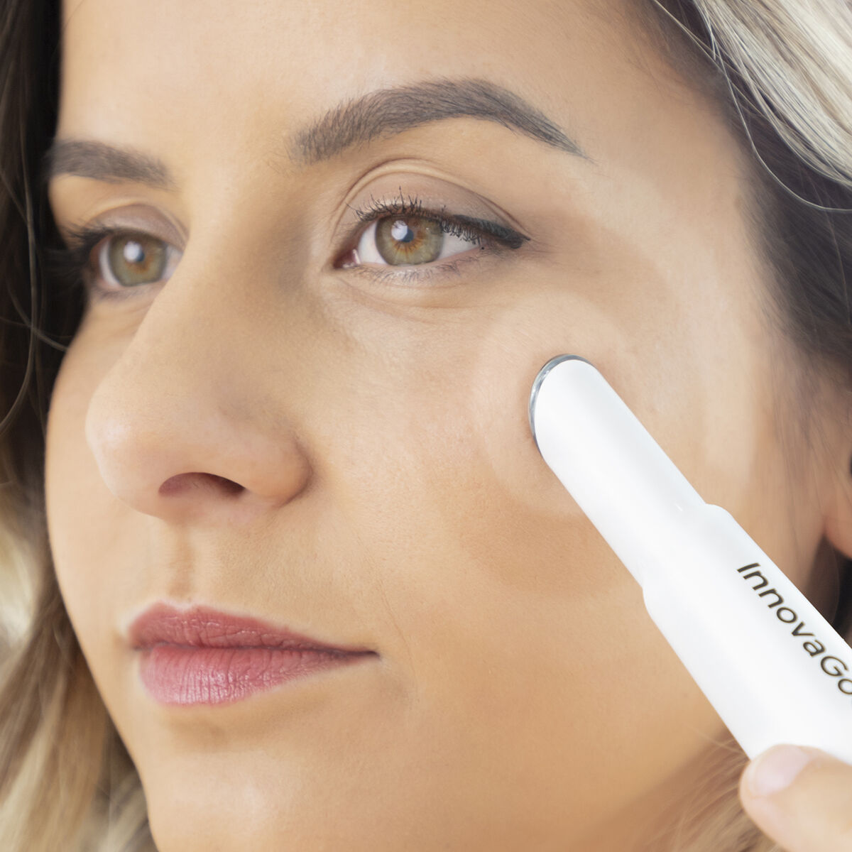 Eine Frau benutzt ein weißes InnovaGoods-Schönheitsgerät unter ihrem Auge. Sie hat helle Haut, braune Augen und blonde Strähnchen.