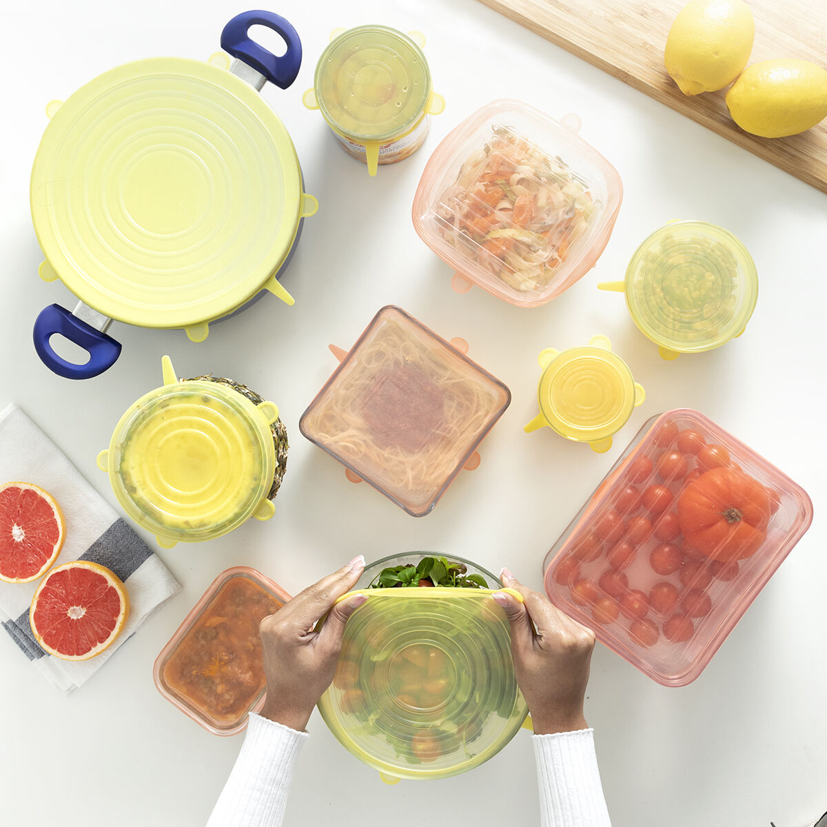 Vari contenitori per alimenti con coperchi, tra cui pomodori, insalata e frutta, sono disposti su una superficie bianca. Mani che mettono un coperchio.