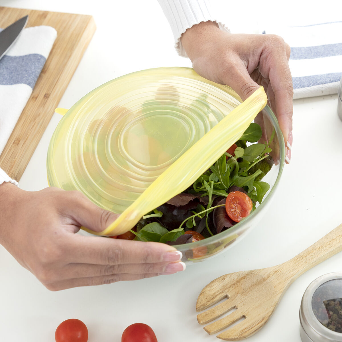 Mani che posizionano un coperchio in silicone giallo su una ciotola di vetro piena di insalata. Si vedono pomodorini e verdure.