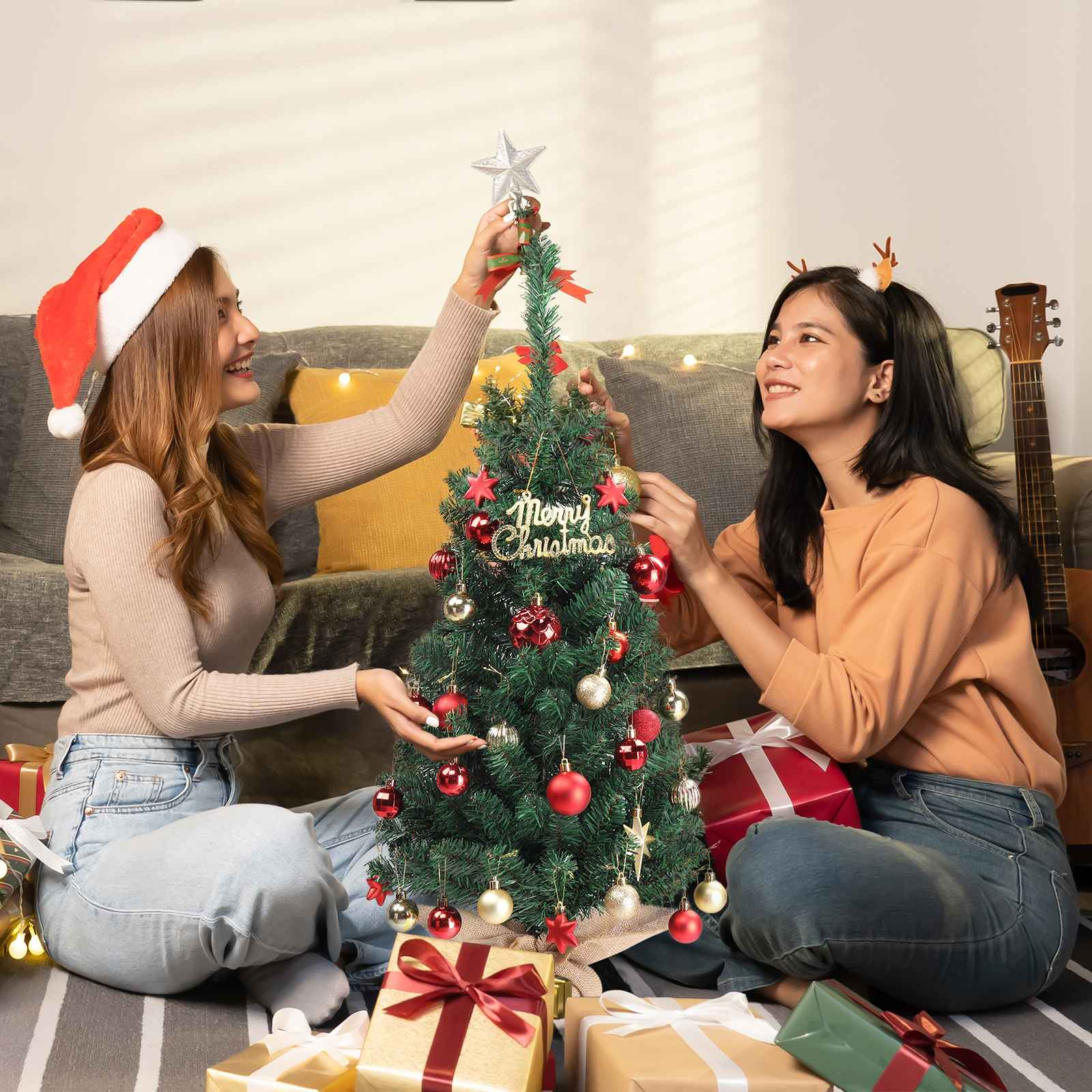 Zwei Frauen schmücken einen kleinen Weihnachtsbaum. Eine trägt einen Weihnachtsmannhut. Geschenke und eine Gitarre sind da.