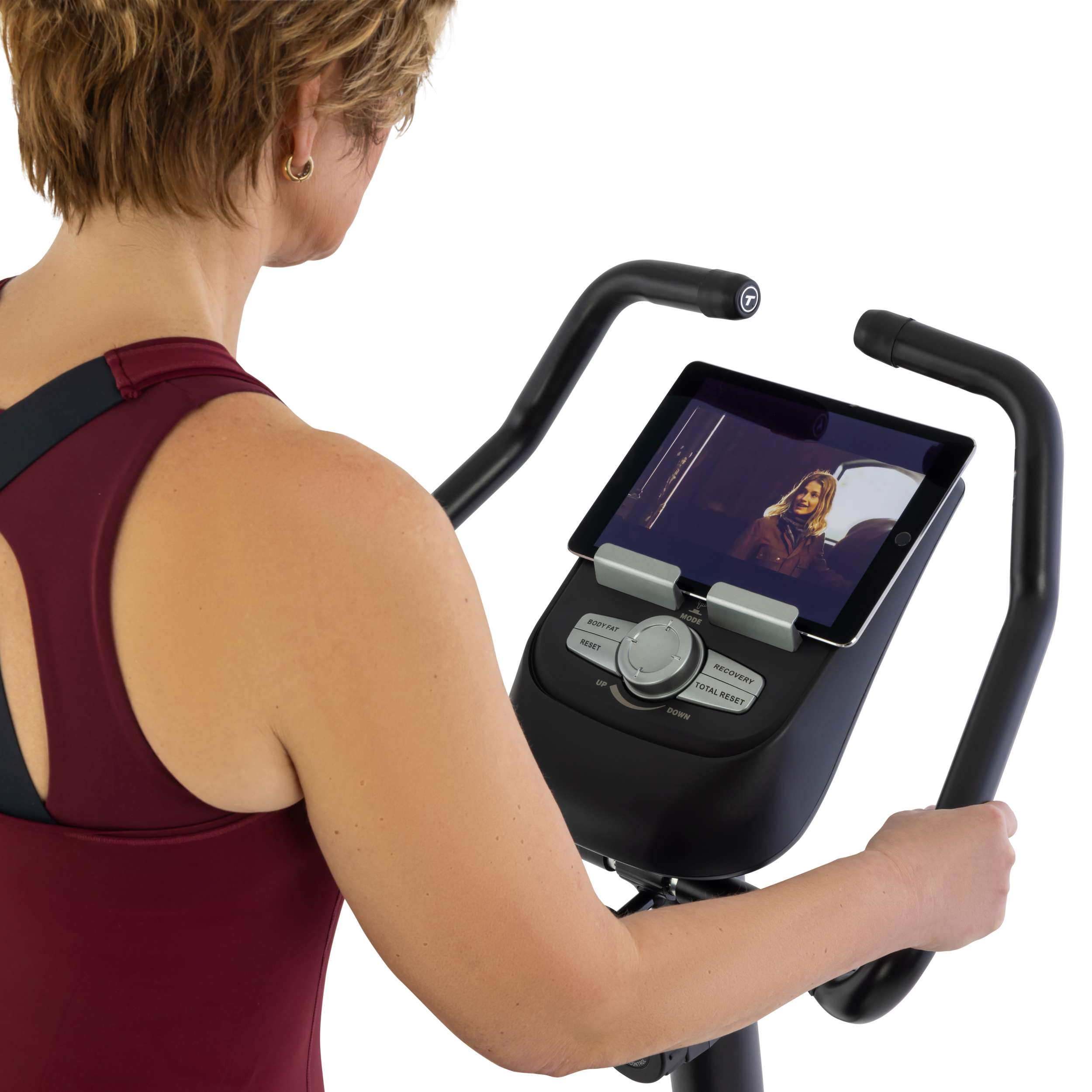 Vrouw op hometrainer. Ze bekijkt een video op een tablet. Ze draagt een bordeauxrode top.