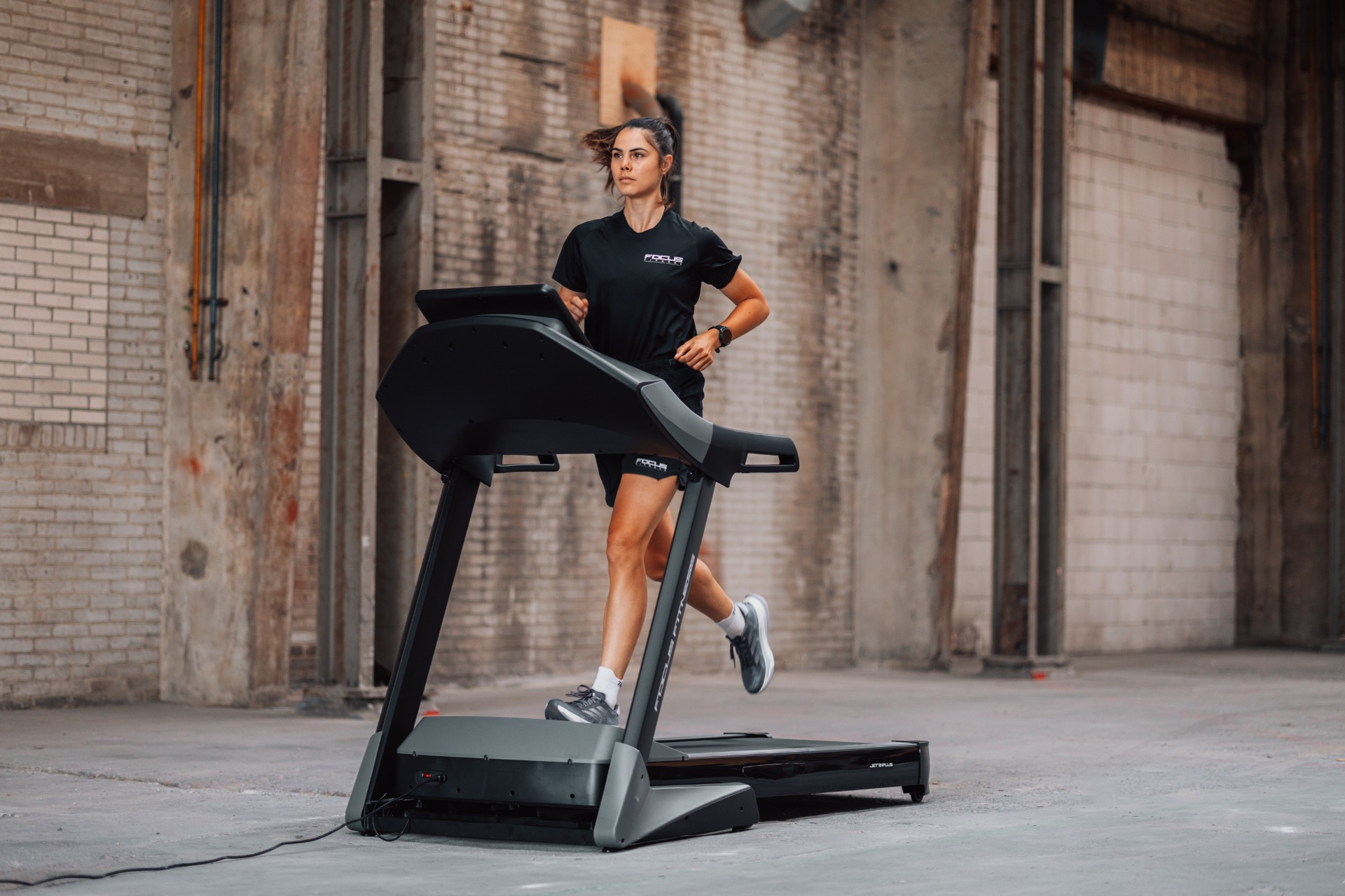A woman runs on a treadmill in an industrial space. She wears black athletic clothes and gray shoes. The background is brick.