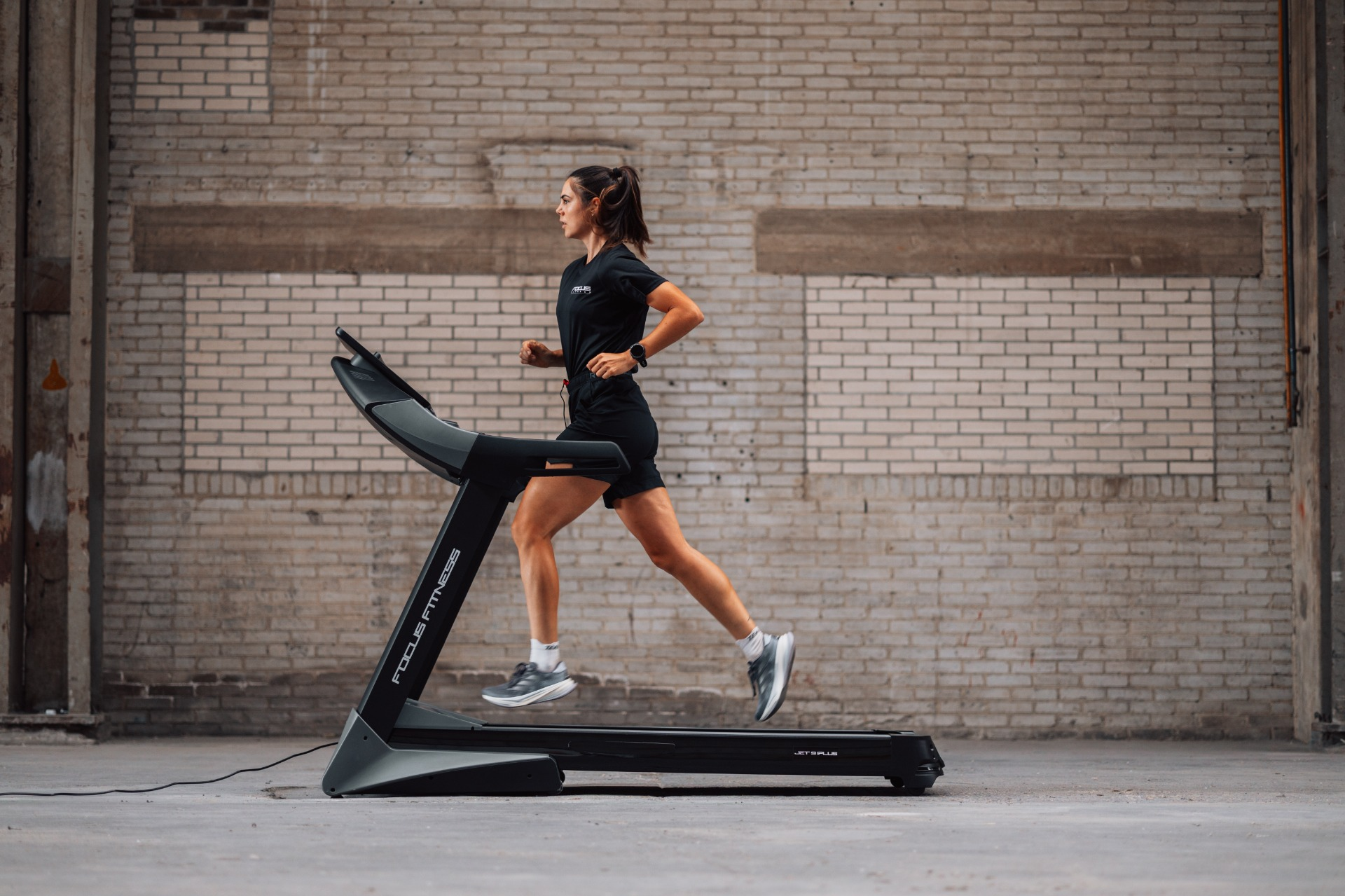 A woman in black athletic wear runs on a treadmill. The background is a brick wall in a large, open space.