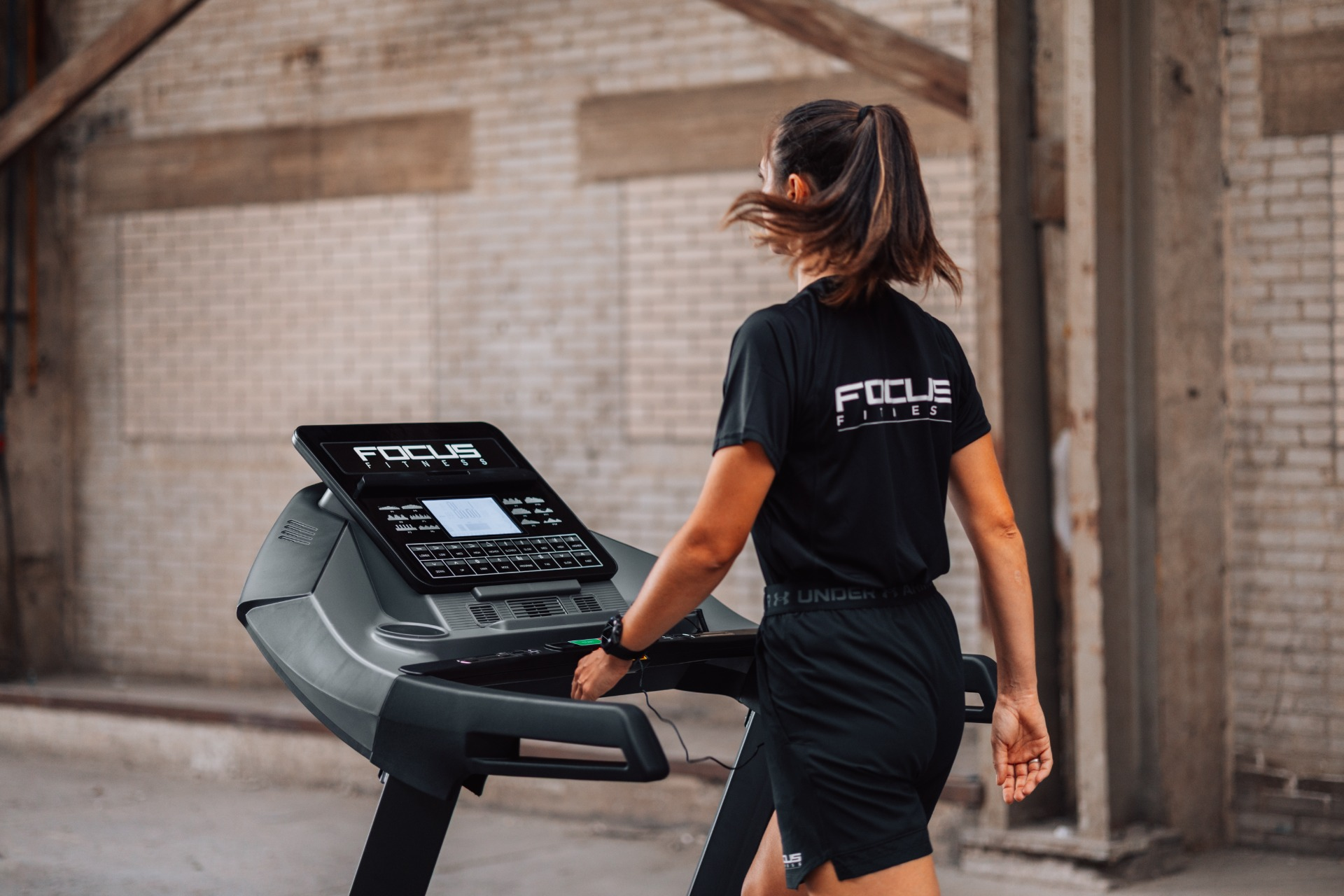 A woman in black workout clothes is on a treadmill. The shirt has the words 'FOCUS FITNESS'. The background is a brick wall.