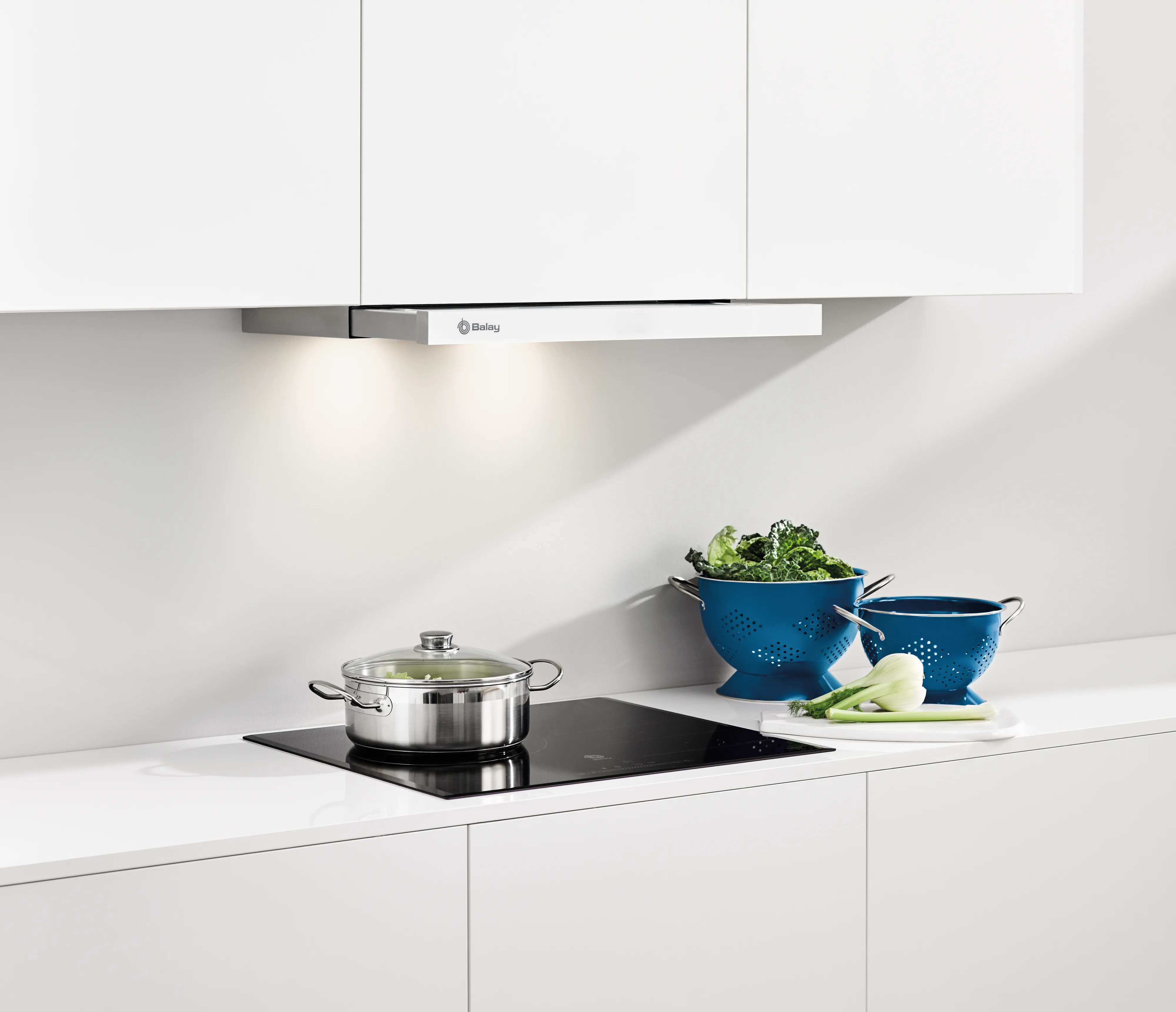 Keuken met pan op fornuis, vergieten en groenten. Witte kasten en afzuigkap zijn zichtbaar.