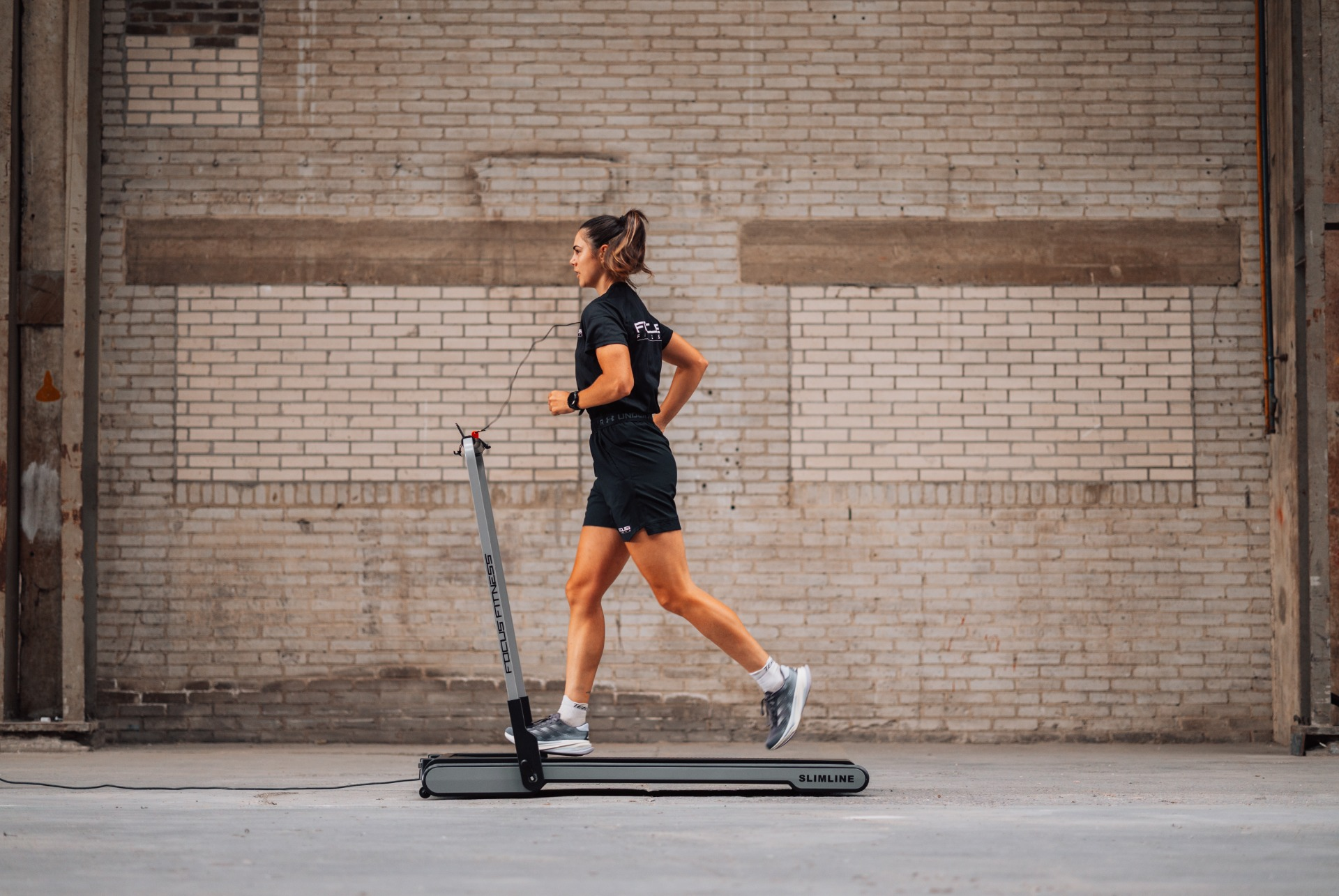 A woman runs on a treadmill. She wears black shorts and shirt. The treadmill is in front of a brick wall.