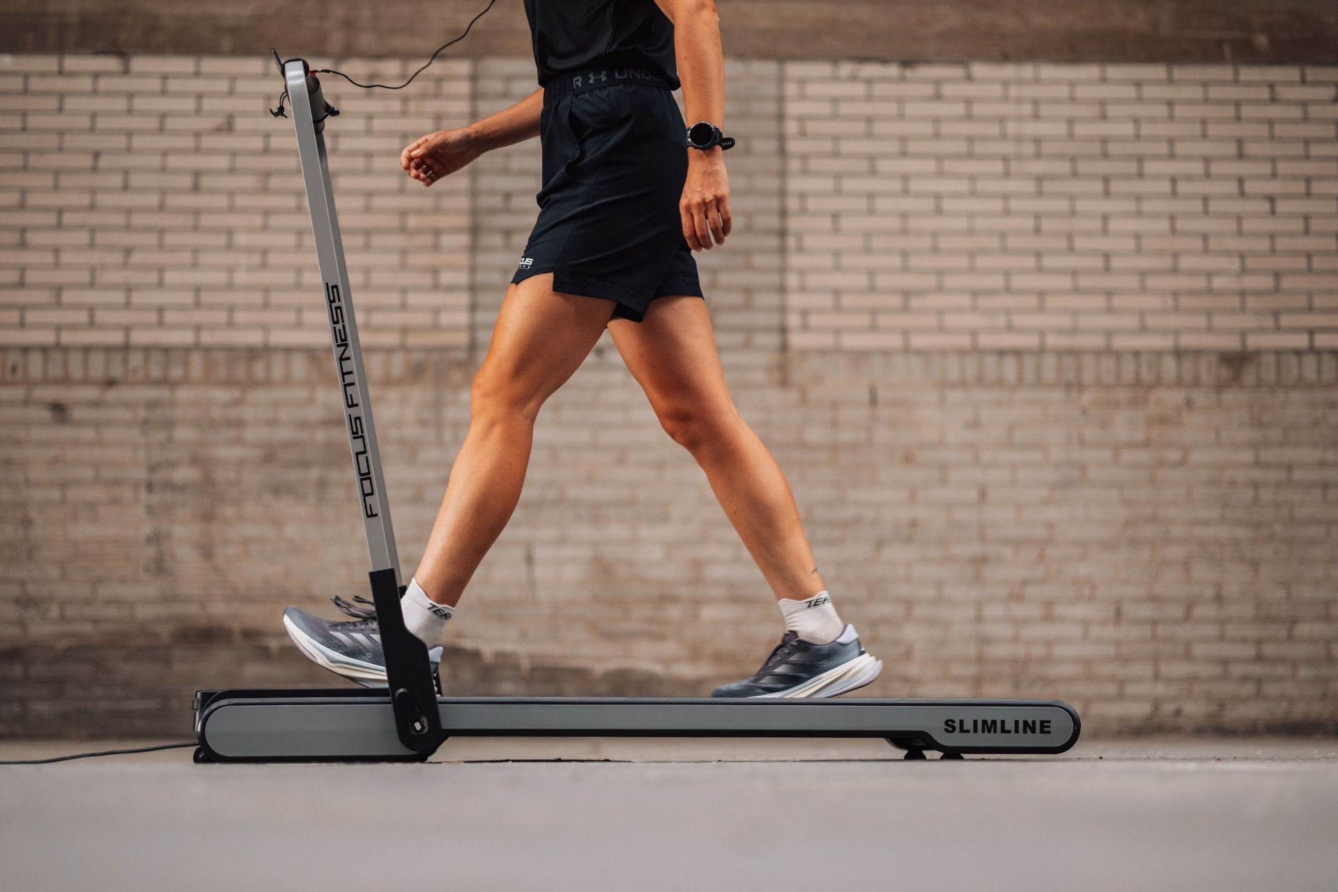 A person walks on a treadmill. They wear black shorts, shoes, and a watch. A brick wall is in the background.