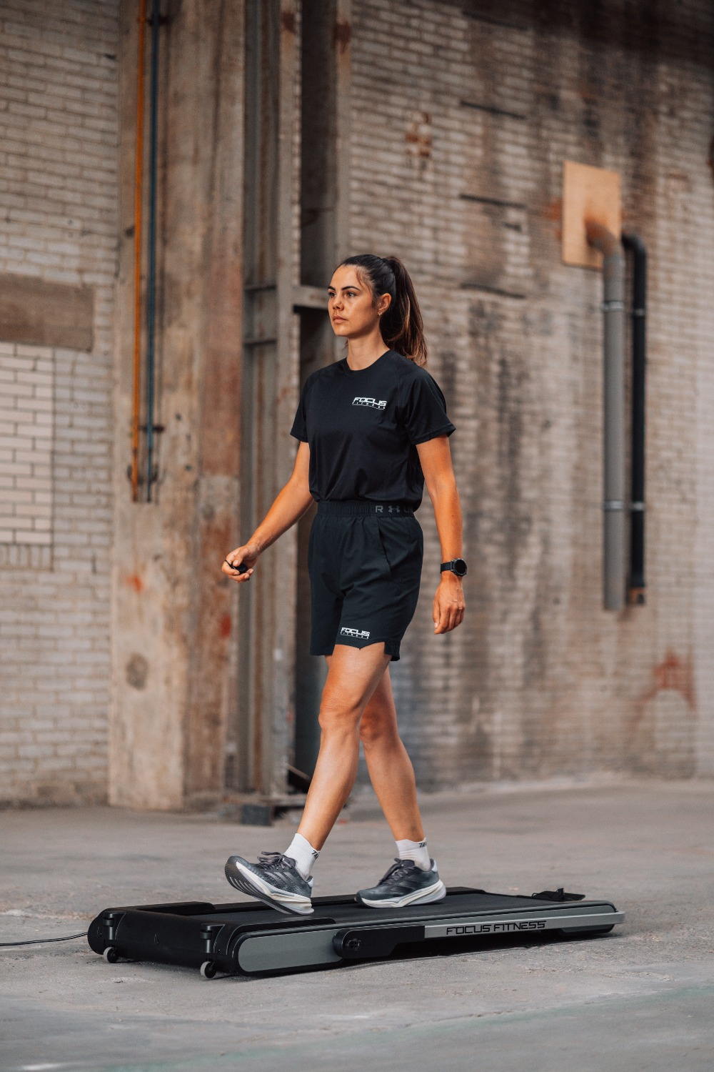 A woman walks on a treadmill indoors. She wears black shorts, t-shirt, and shoes. The background is a brick wall.