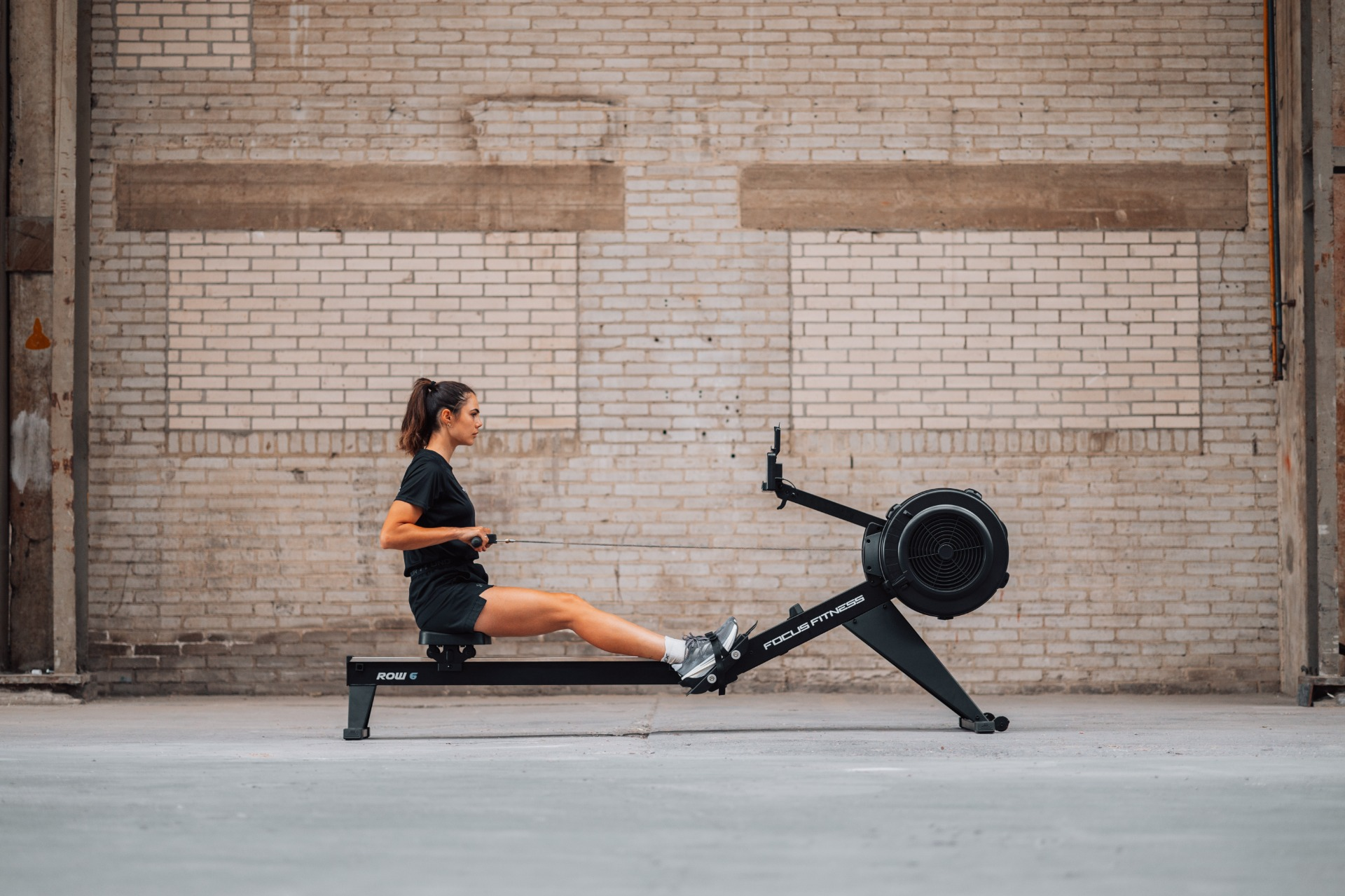 A woman on a rowing machine in a brick-walled room. She's pulling the handles. The machine says 'Focus Fitness'.