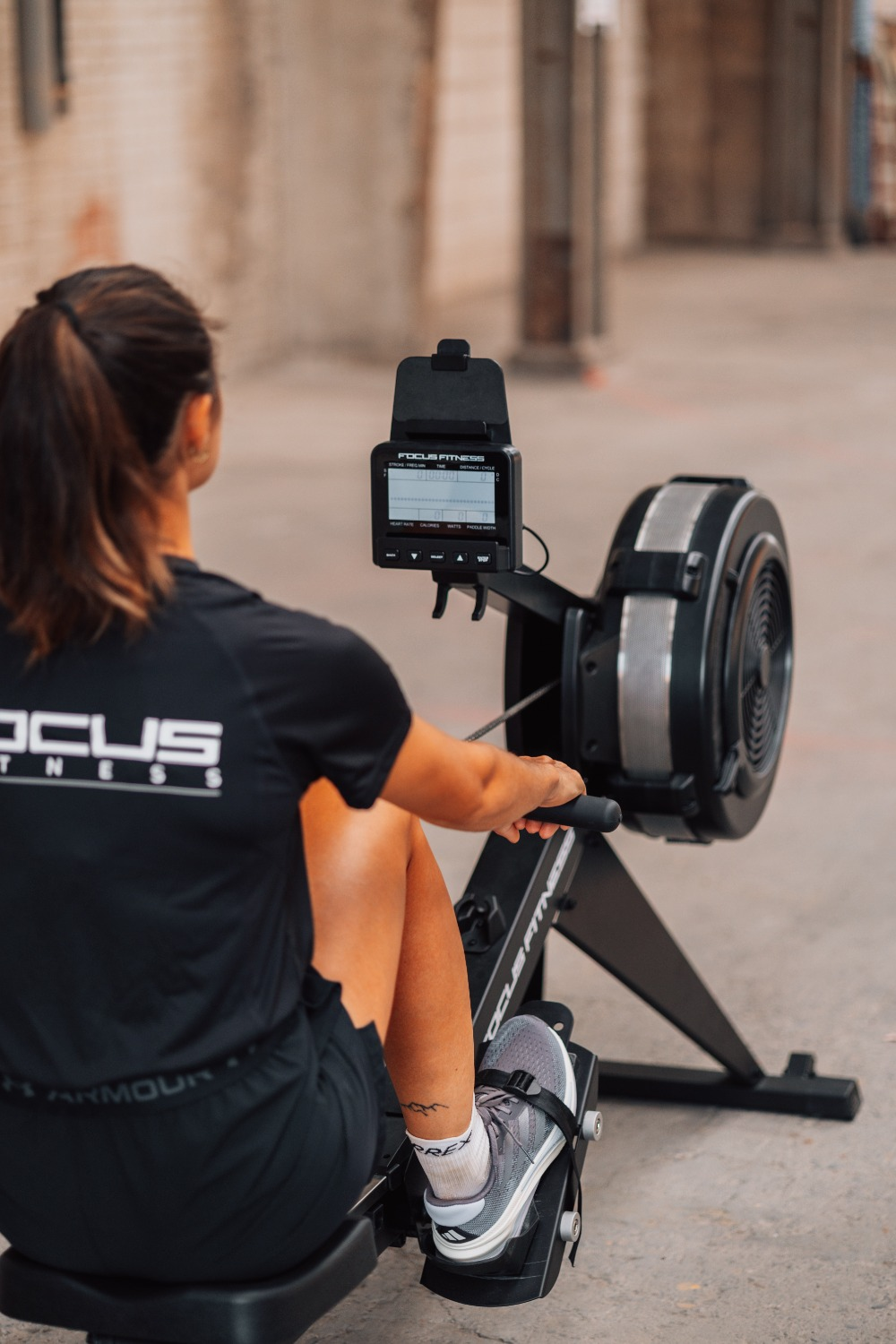 A woman is using a rowing machine. She is wearing a black shirt and shorts. The machine has a display showing workout stats.