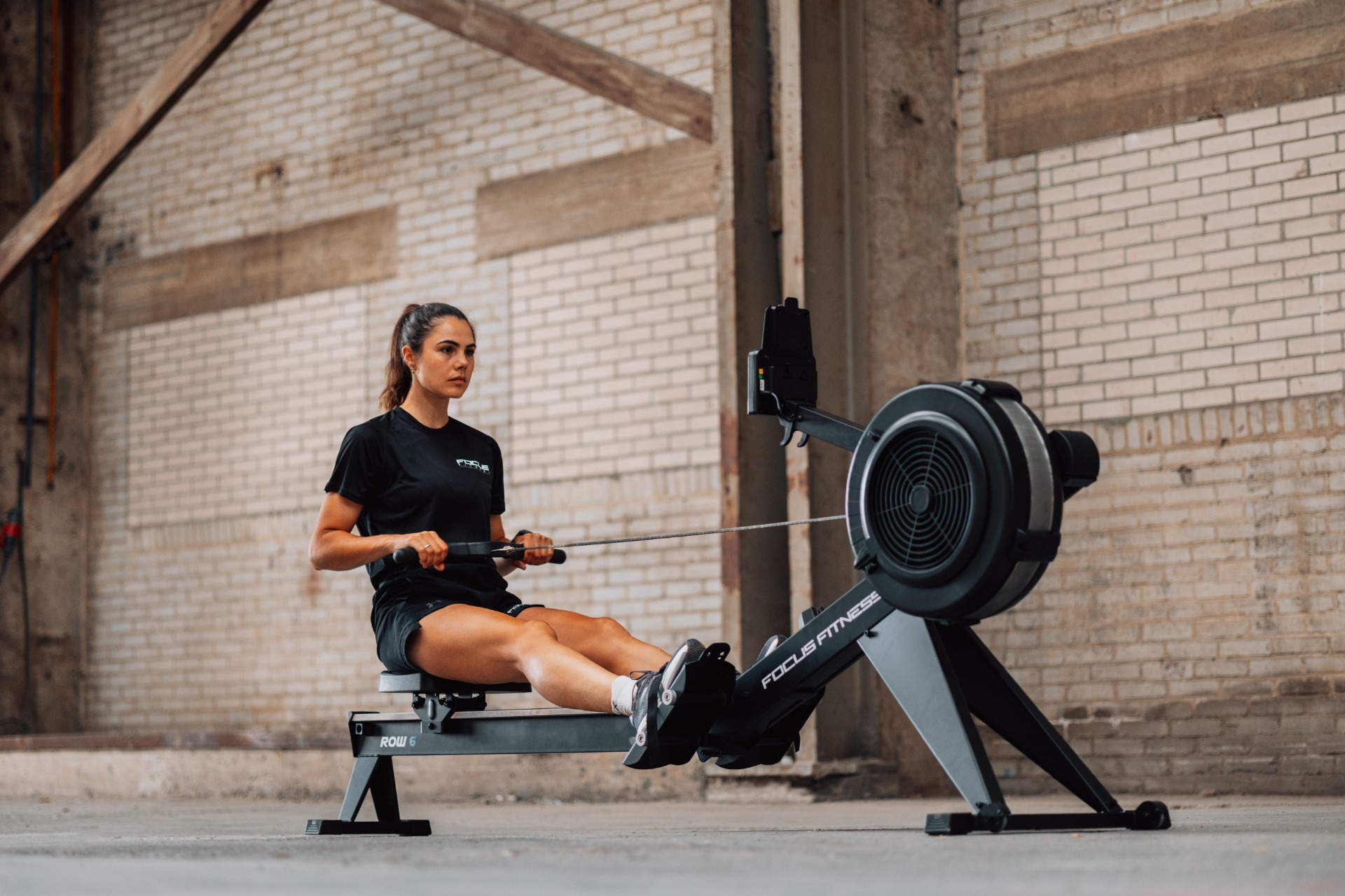 A woman is rowing on a Focus Fitness machine in a brick-walled gym. She wears black athletic wear.