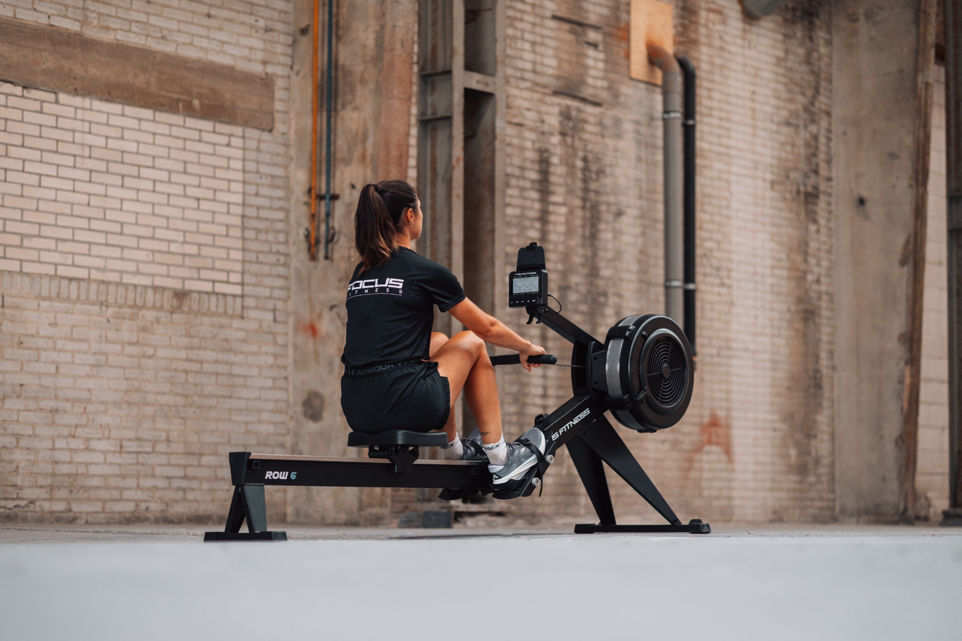 A woman is rowing on a black rowing machine. She wears black shorts and a t-shirt. The background is a brick wall.