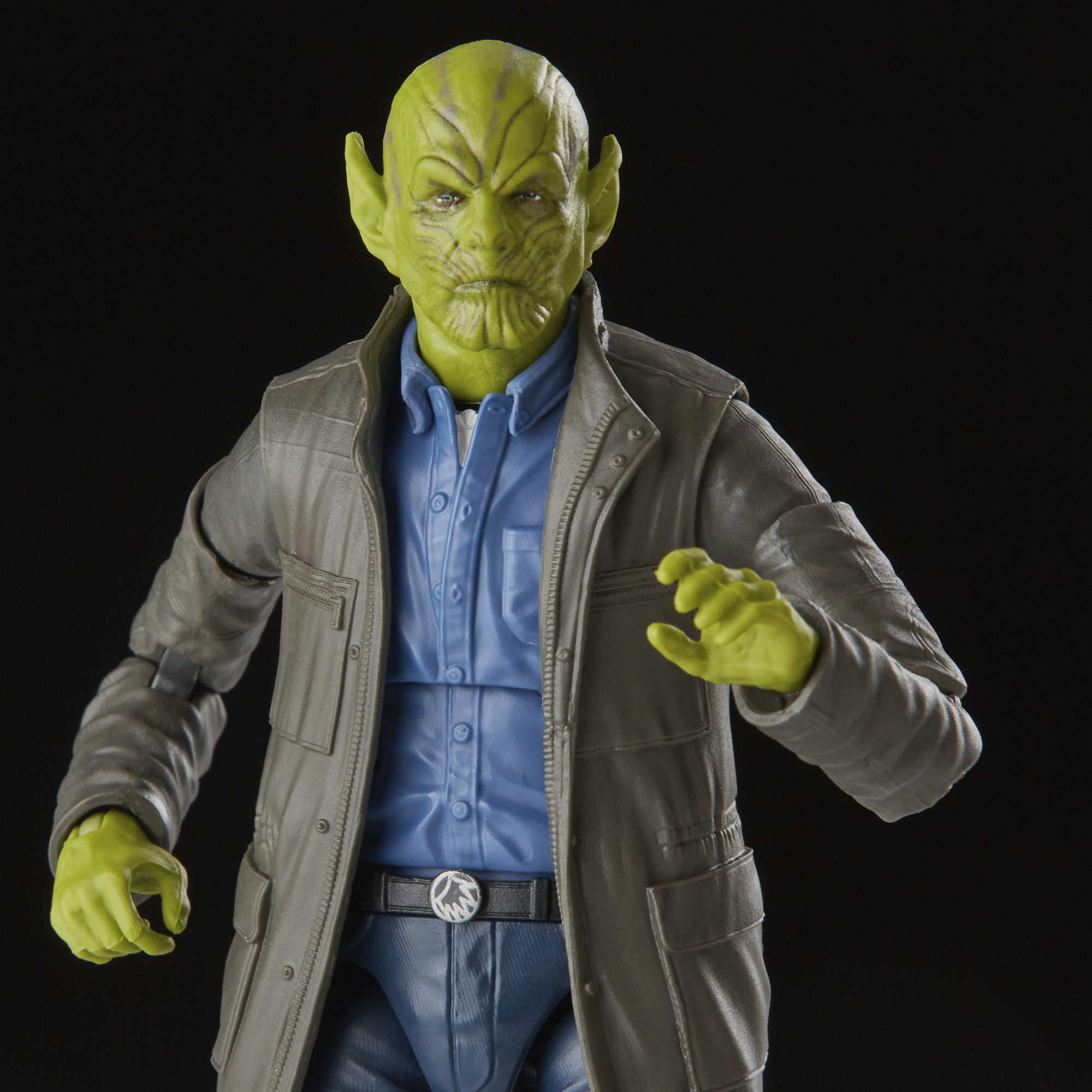 Una figura de piel verde con orejas grandes, que lleva una chaqueta gris sobre una camisa y pantalones azules. Fondo negro.