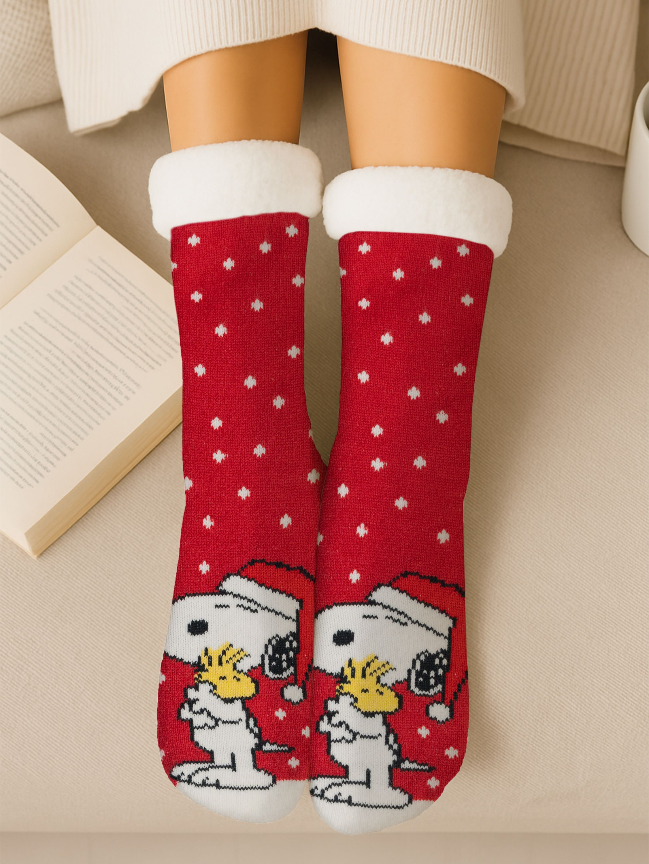 Person trägt rote Socken mit Snoopy- und Woodstock-Designs, weißen Verzierungen und weißen Tupfen. Im Hintergrund ist ein offenes Buch.