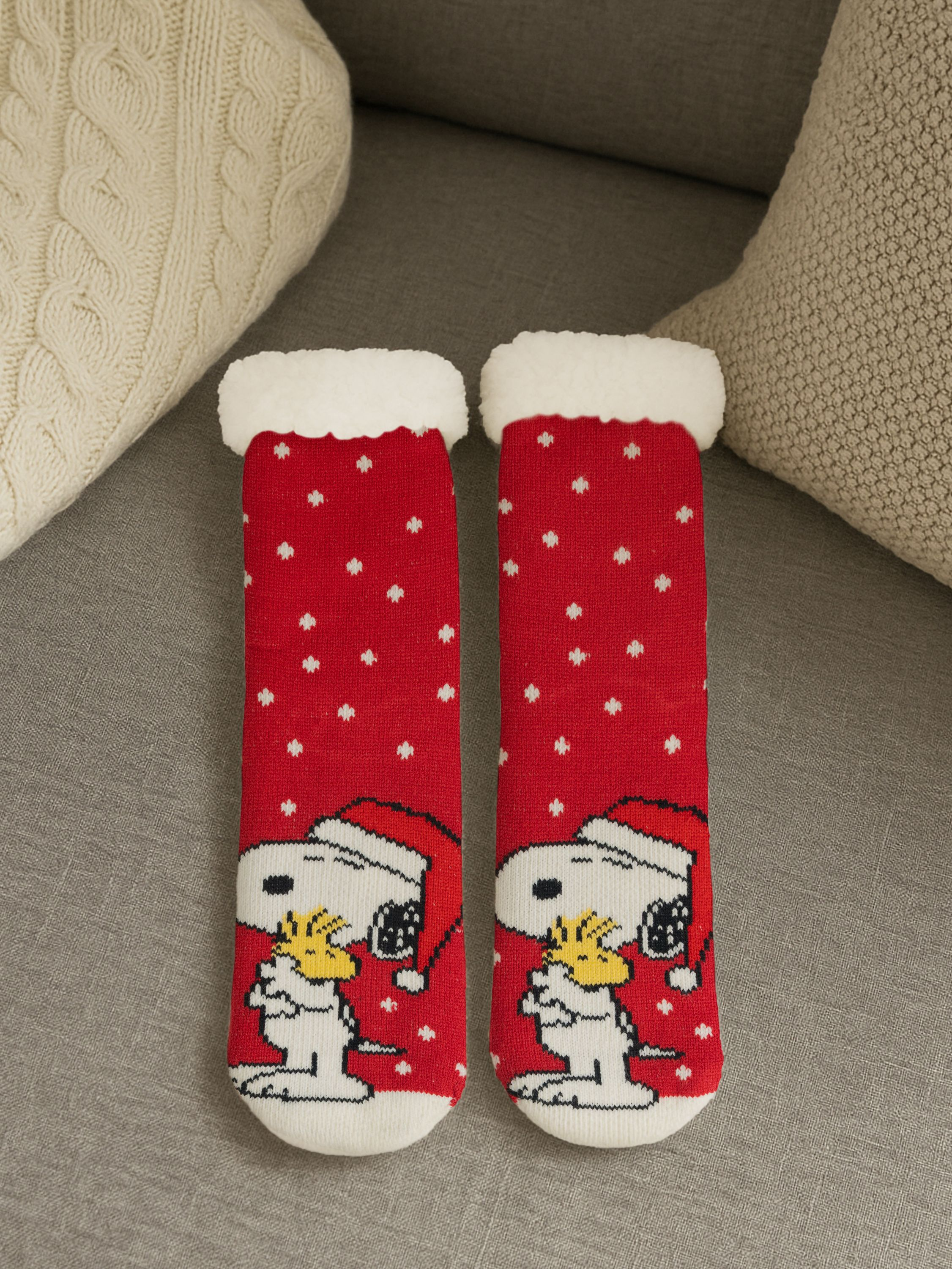 Zwei rote Socken mit Snoopy-Design, weißen Tupfen und weißen, flauschigen Oberteilen. Sie liegen auf einem grauen Sofa.