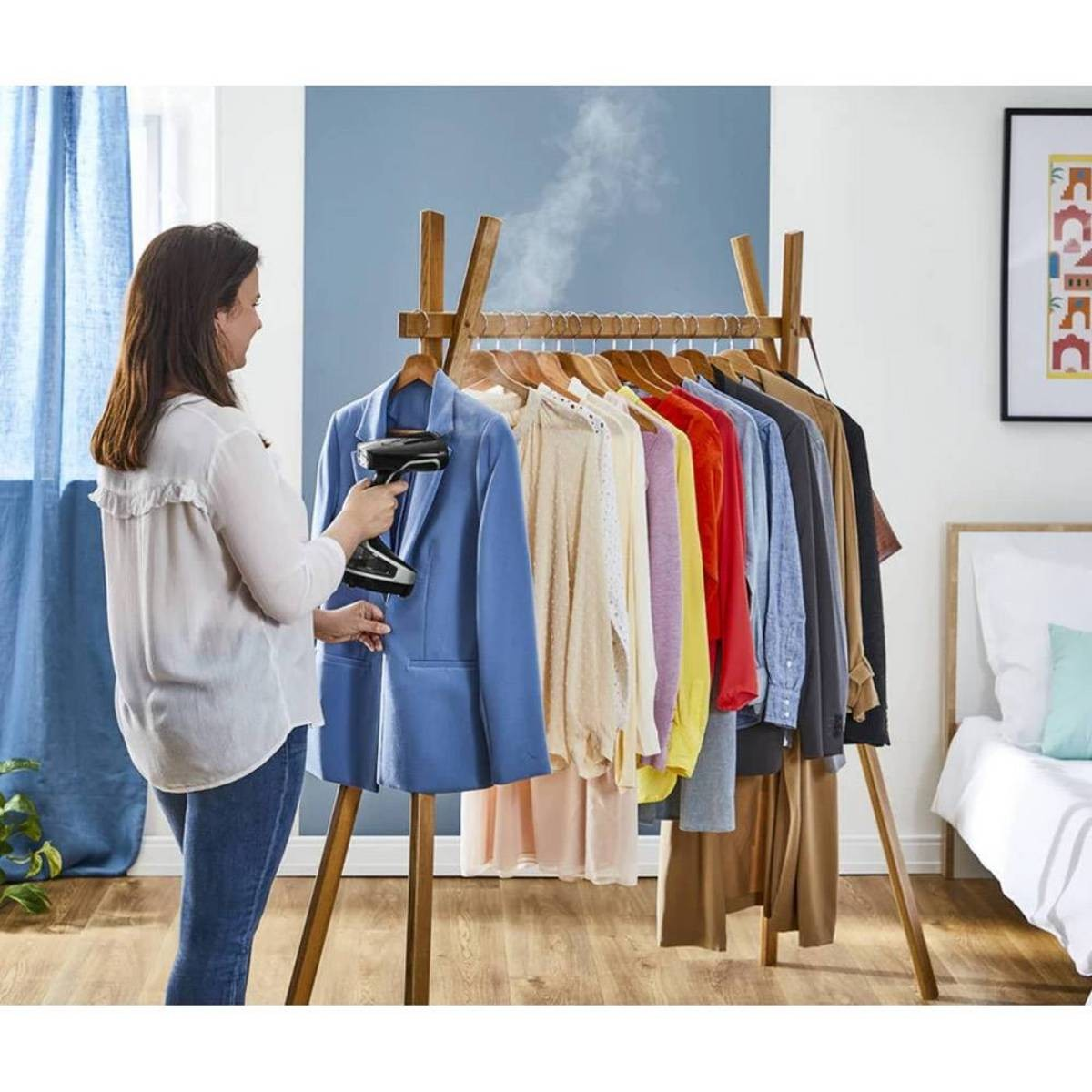 Une femme repasse un blazer bleu sur un portant avec des vêtements de couleurs variées. La vapeur monte. Un lit et une œuvre d'art sont en arrière-plan.