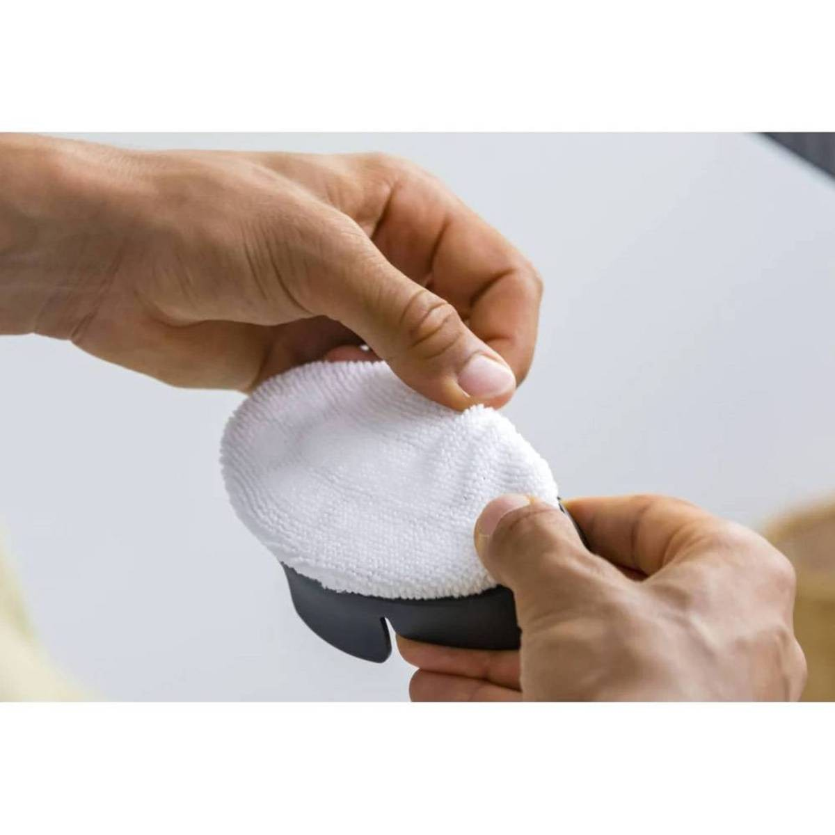 Mains tenant un tampon blanc, rond et duveteux, fixé à une base en plastique gris foncé, probablement pour le nettoyage.