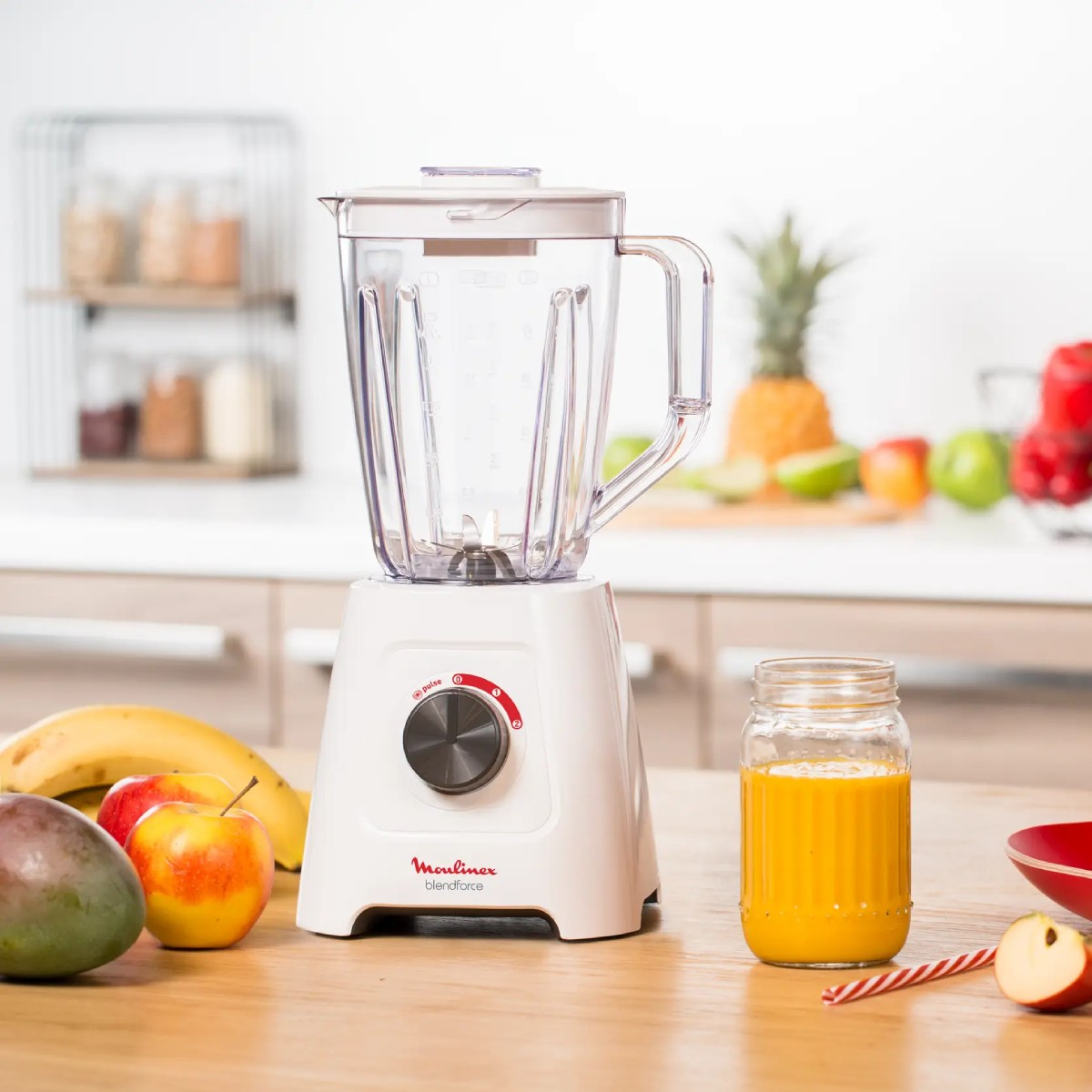Un frullatore bianco con frutta su un tavolo di legno. I frutti sono una banana, una mela e un mango e un barattolo con succo d'arancia.
