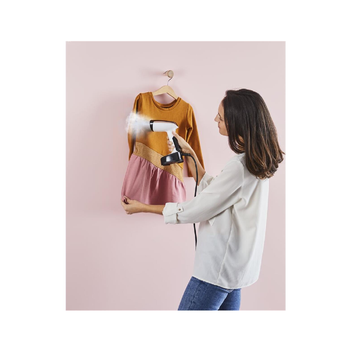 Una mujer plancha un vestido en una percha. El vestido es mostaza y rosa. El fondo es una pared rosa.