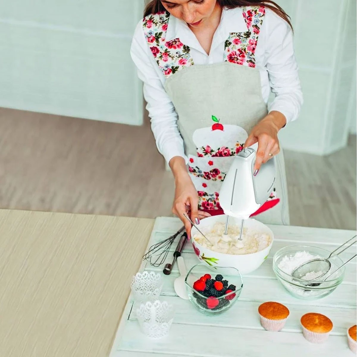 Eine Frau in einer Schürze verrührt Zutaten in einer Schüssel mit einem Mixer. Beeren, Cupcakes und Puderzucker stehen in der Nähe auf dem Tisch.