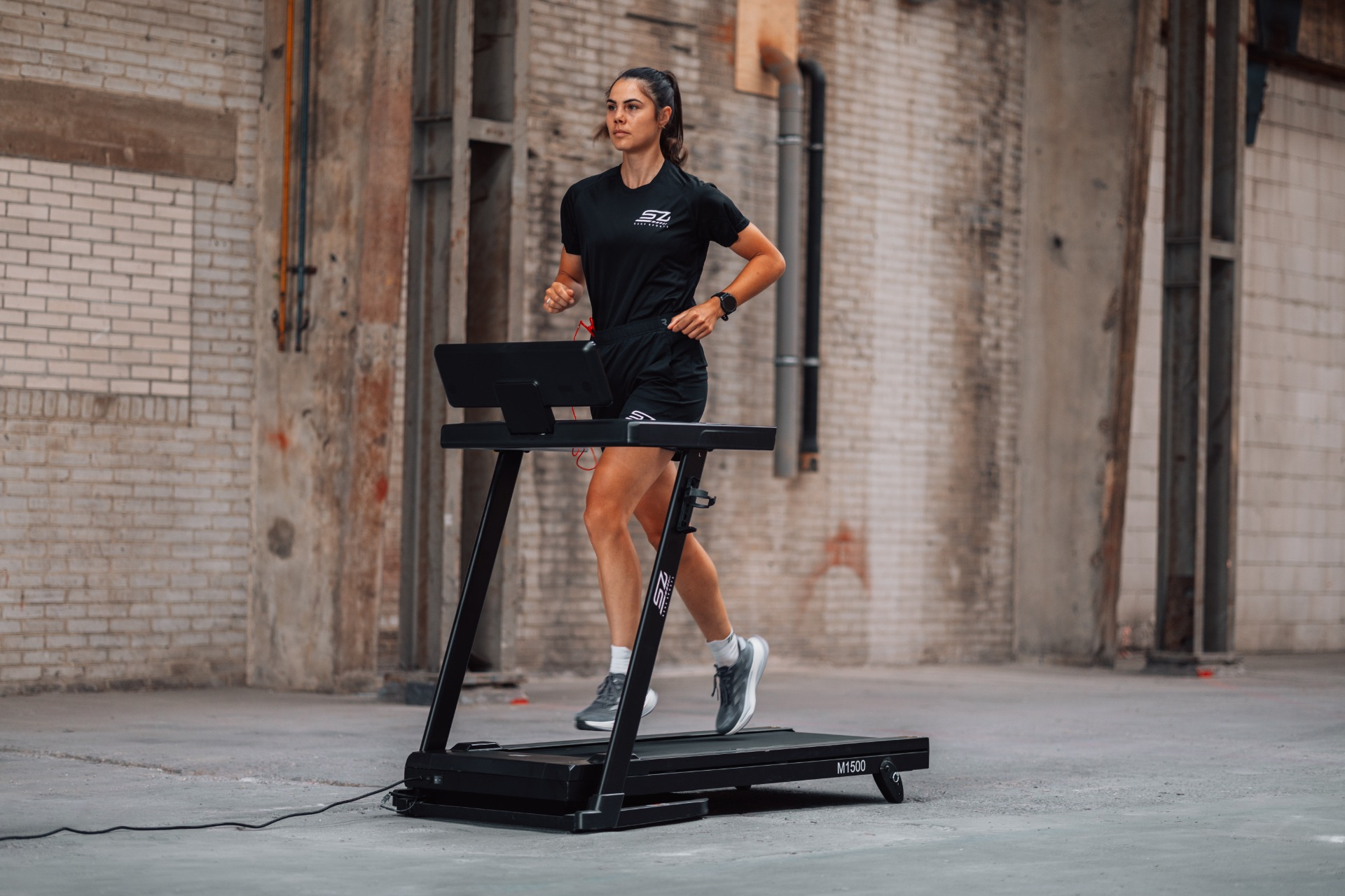 A woman runs on a treadmill in an industrial space. She wears black athletic attire. The background is brick and concrete.