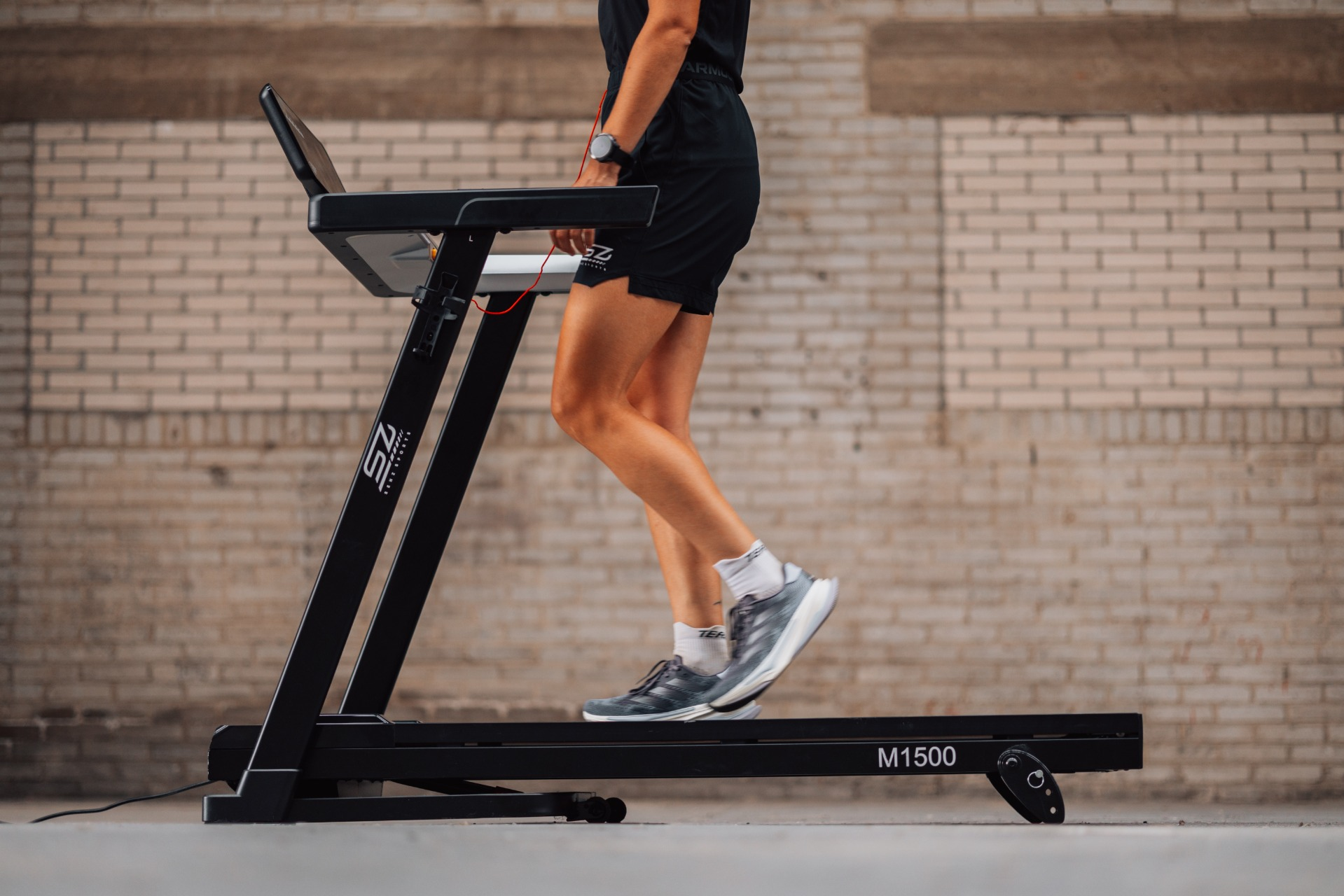 A person runs on a treadmill. The machine is black, and the background is a brick wall. The person wears shorts and a watch.