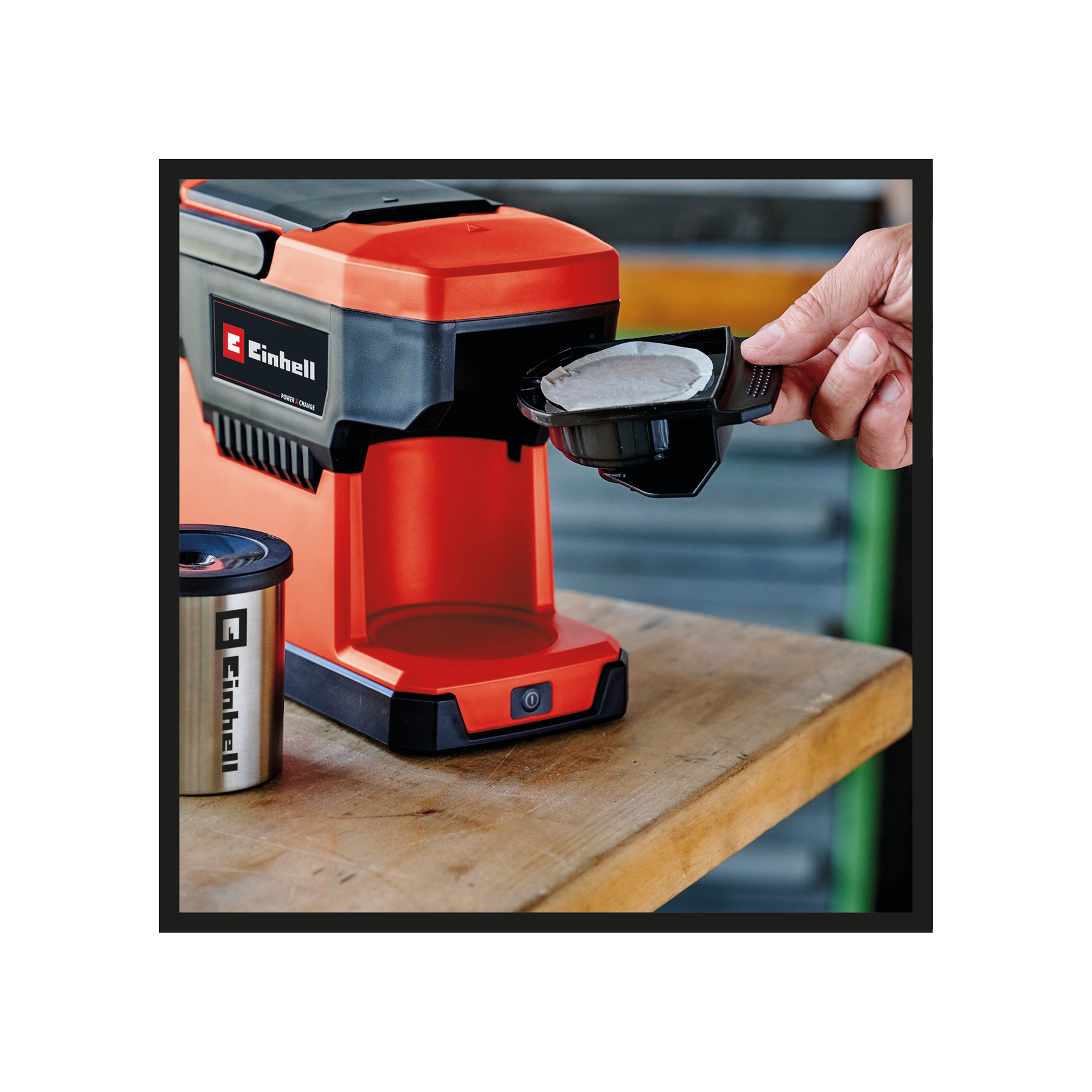 Osoba wkłada kapsułkę kawy do czerwono-czarnego ekspresu do kawy, srebrny kubek stoi na drewnianym stole.