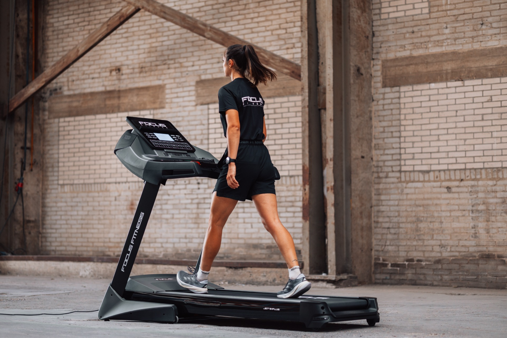A woman in black activewear walks on a treadmill. The background features a brick wall and wooden beams.