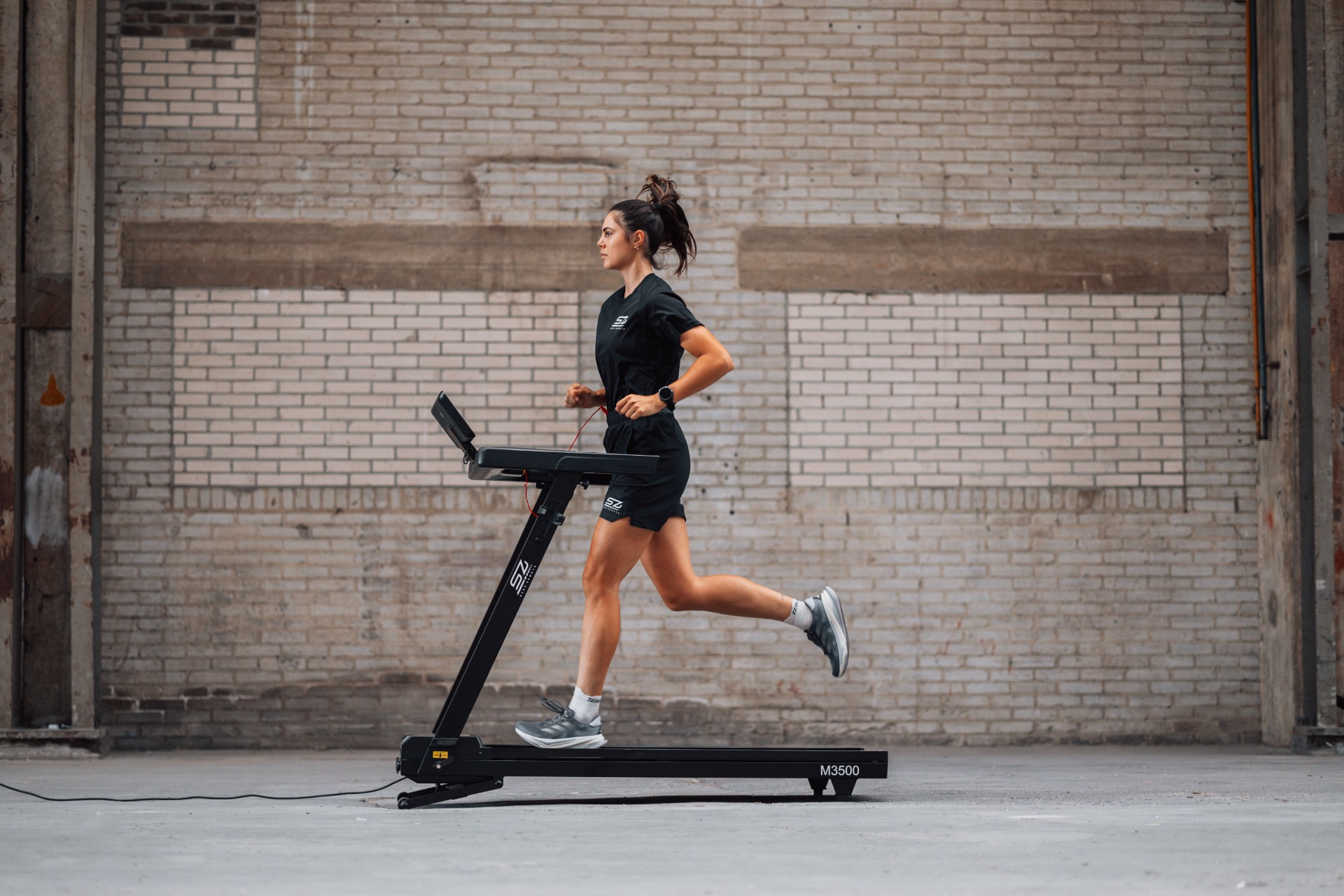 A woman runs on a treadmill in a brick-walled room. She wears black shorts and a shirt. The machine is black.