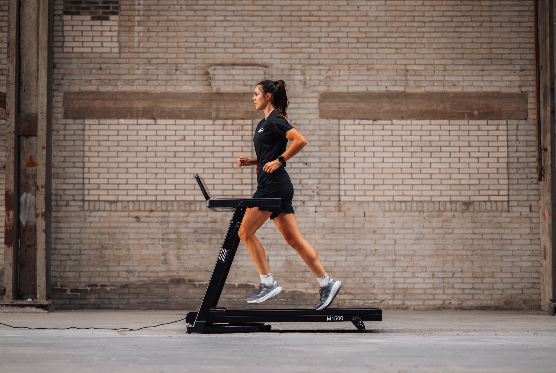 A woman runs on a treadmill in a warehouse. She wears black athletic wear. The background is a brick wall.