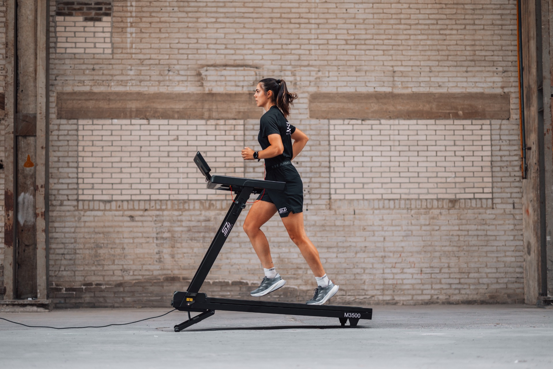 A woman runs on a treadmill in a brick-walled room. She wears black athletic wear and gray shoes. The treadmill is black.