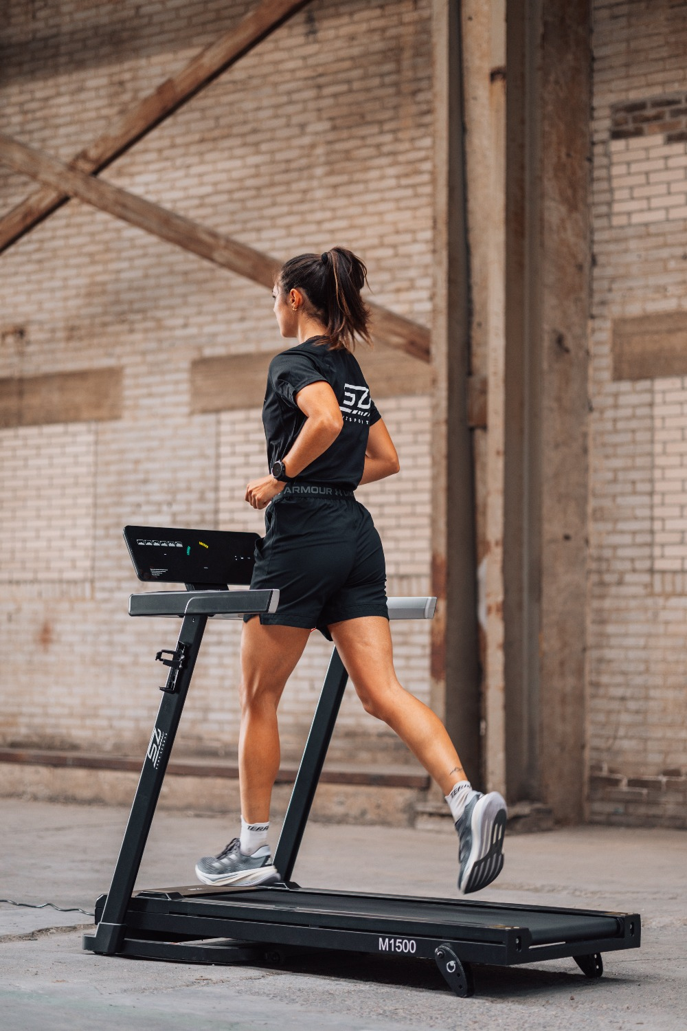Woman runs on a treadmill in a brick-walled industrial space. She wears black athletic wear and gray sneakers. The treadmill is black.