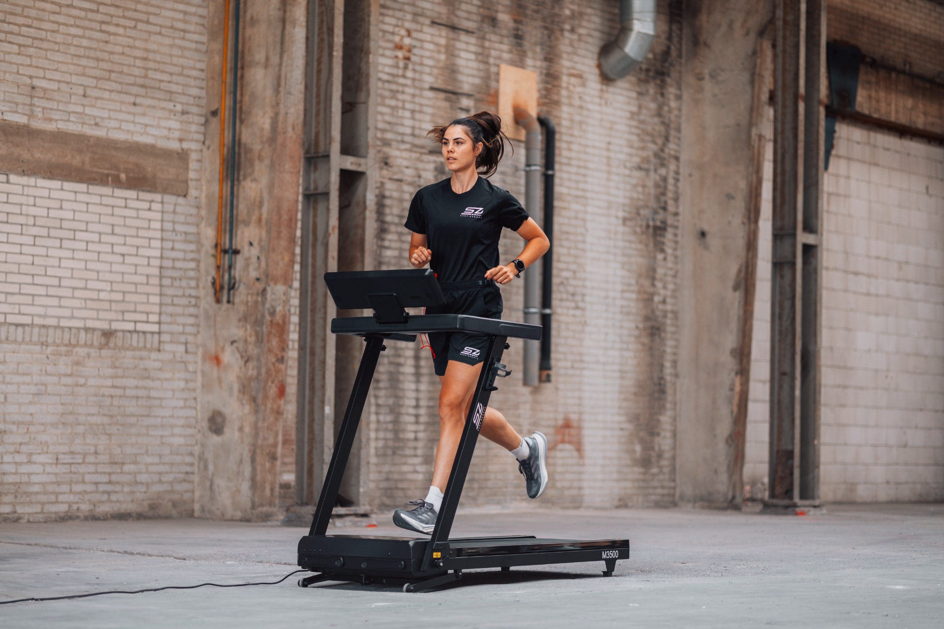 Woman running on a treadmill in an industrial space. She wears black athletic wear. The treadmill is black.