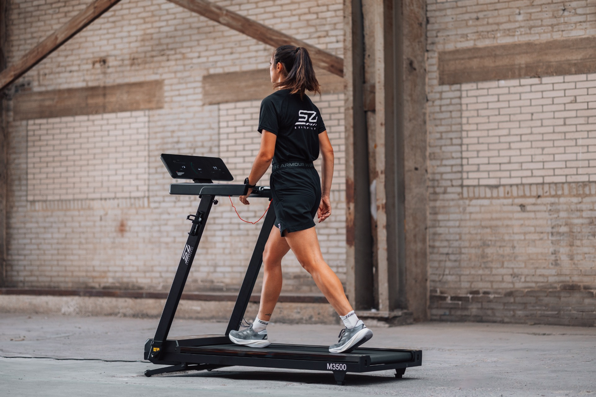 A woman walks on a treadmill in a brick-walled space. She wears black shorts and a tee. The treadmill is black.