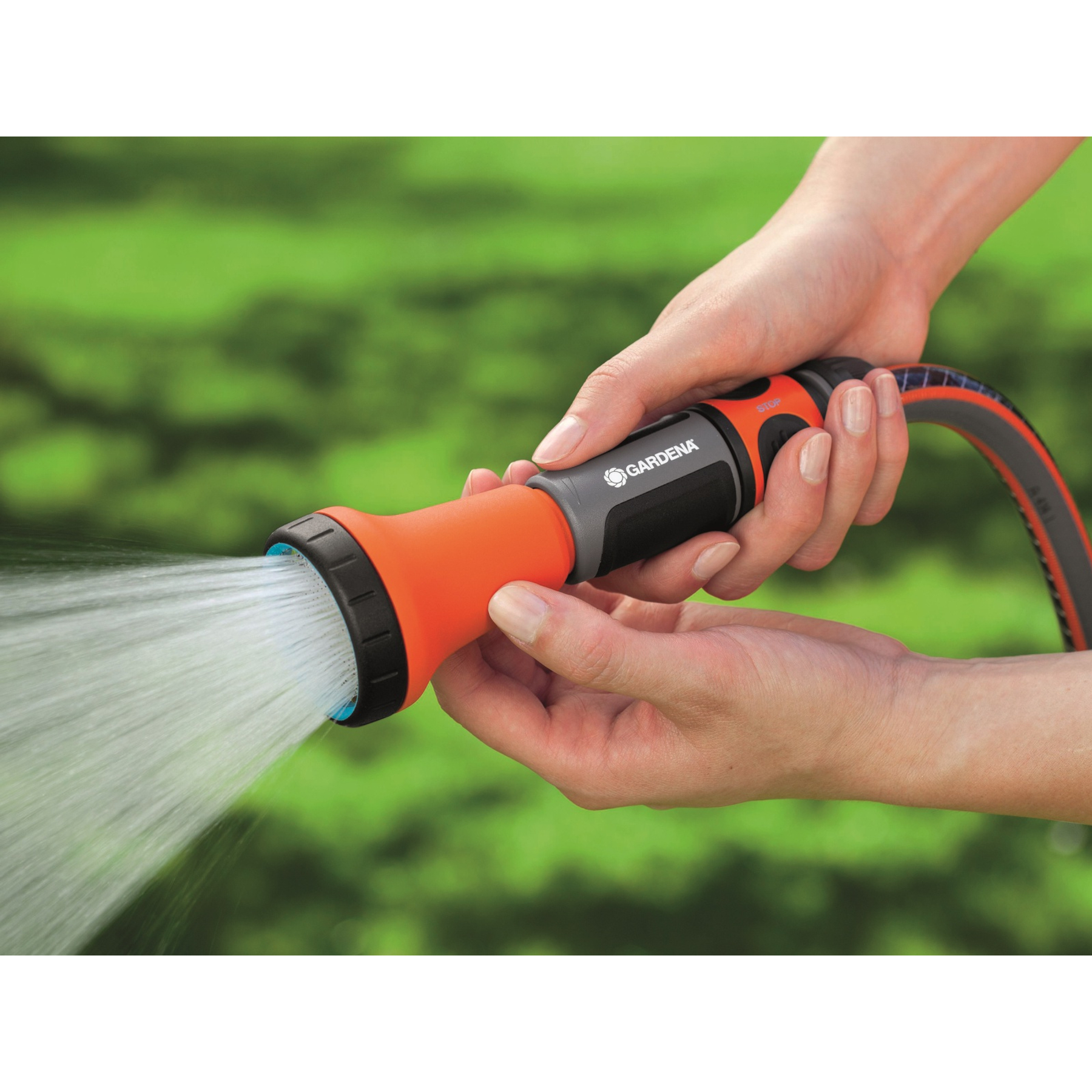 Le mani tengono un ugello per l'acqua arancione Gardena, che spruzza acqua. Sfondo verde sfocato.