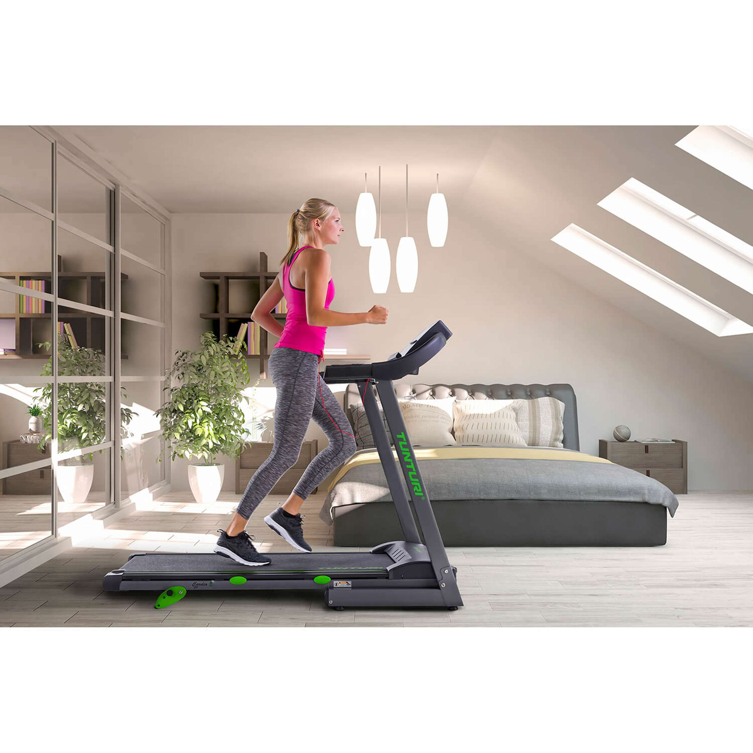 Een vrouw rent op een loopband in een lichte kamer. Een bed en planten zijn zichtbaar. Boven, hanglampen.