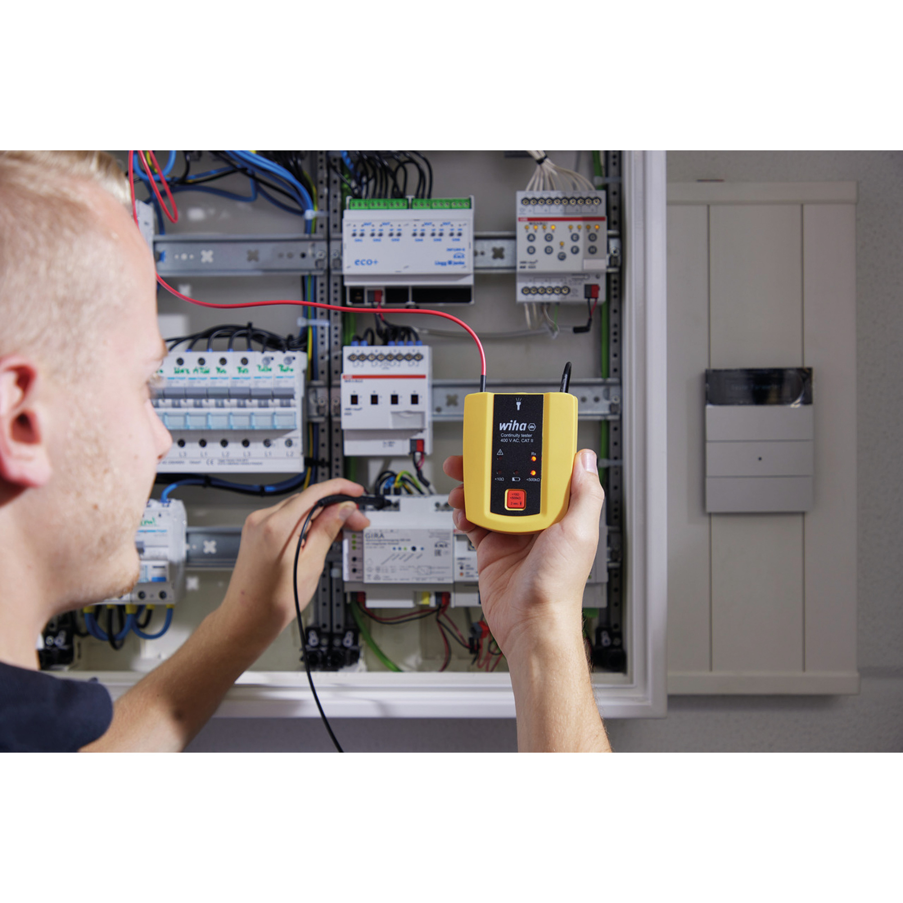 Ein Mann testet elektrische Schaltkreise mit einem gelben Wiha-Tester in einem offenen Schaltschrank. Drähte und Komponenten sind sichtbar.
