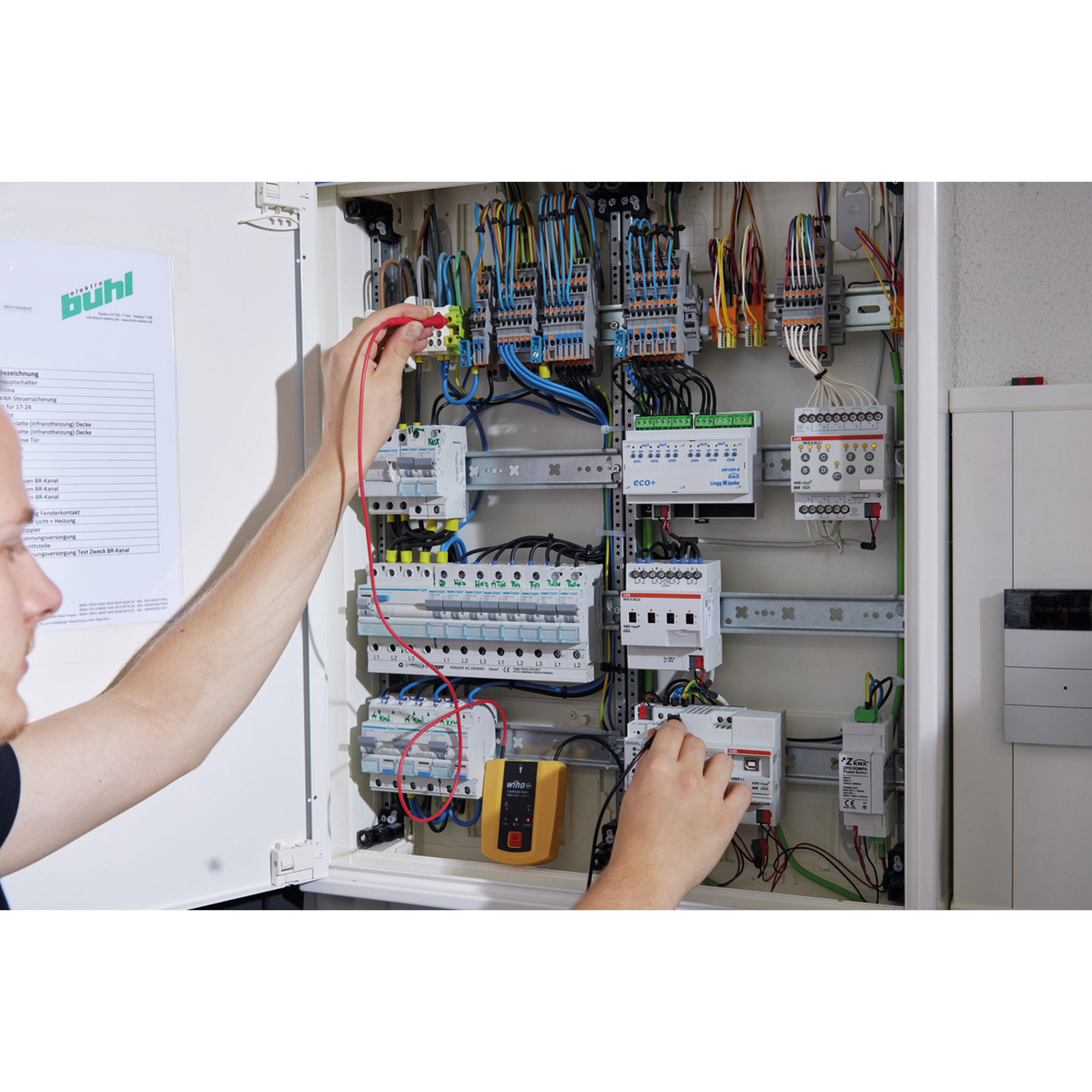 Ein Elektriker testet Drähte in einer Schalttafel. Kabel und Komponenten sind sichtbar. Ein gelbes Gerät wird gehalten.