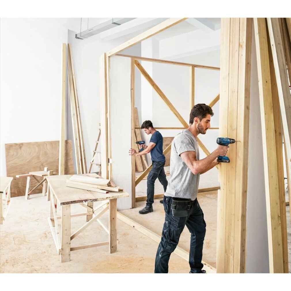 Dwóch mężczyzn używa wiertarek do montażu drewnianych ram w pomieszczeniu. Deski i inne narzędzia sugerują prace budowlane.