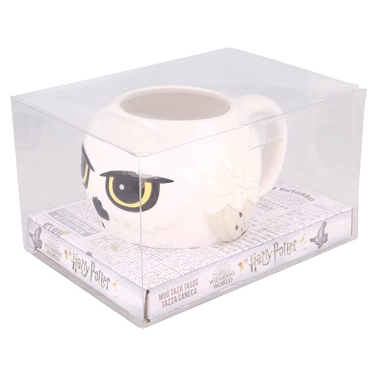 Una taza blanca de Harry Potter con forma de búho está en una caja transparente sobre una caja con el logo de Harry Potter y texto en inglés y otros idiomas.