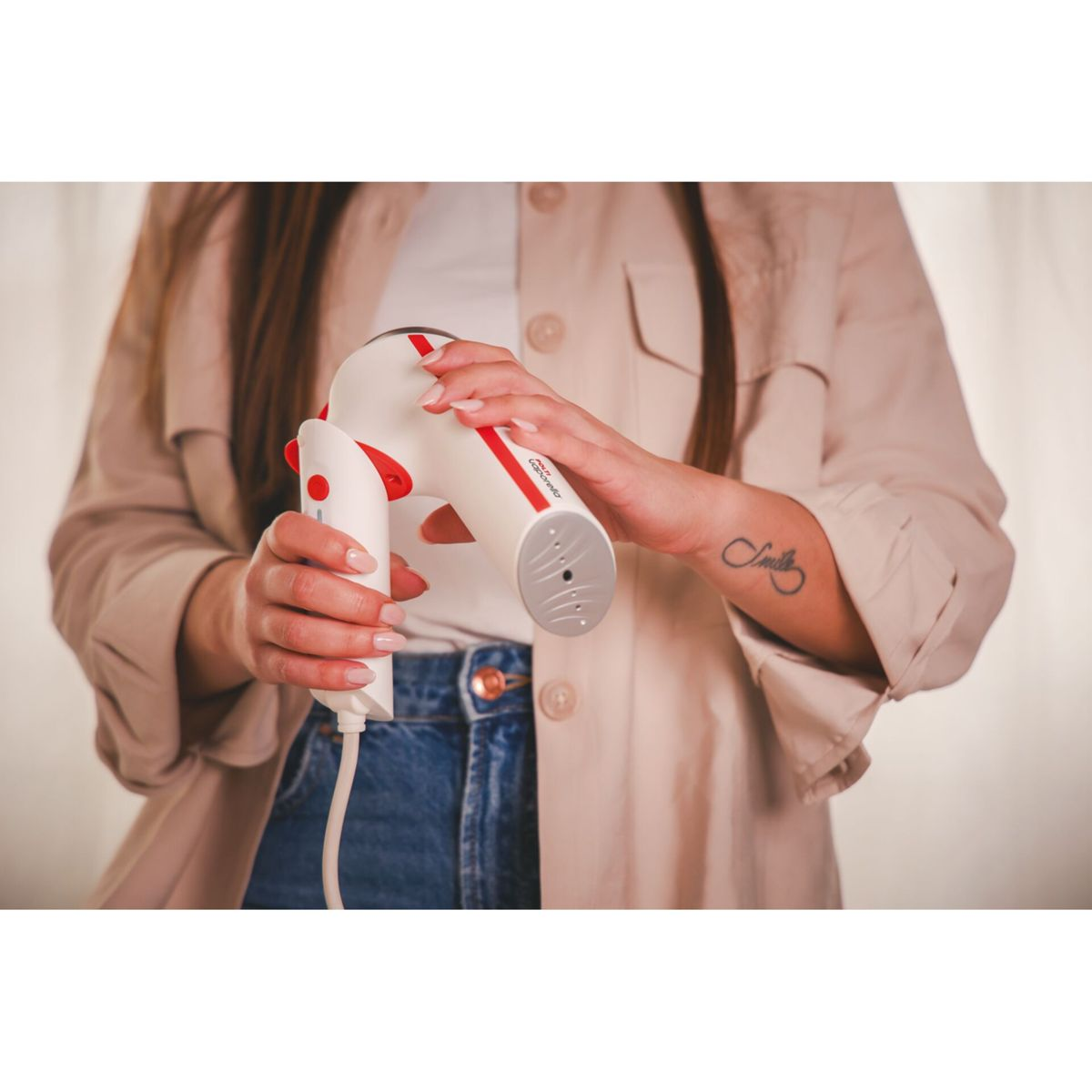 Eine Person hält einen weißen und roten Kleidungsdampfer. Sie trägt eine beige Jacke, blaue Jeans und hat ein Tattoo auf dem Arm.